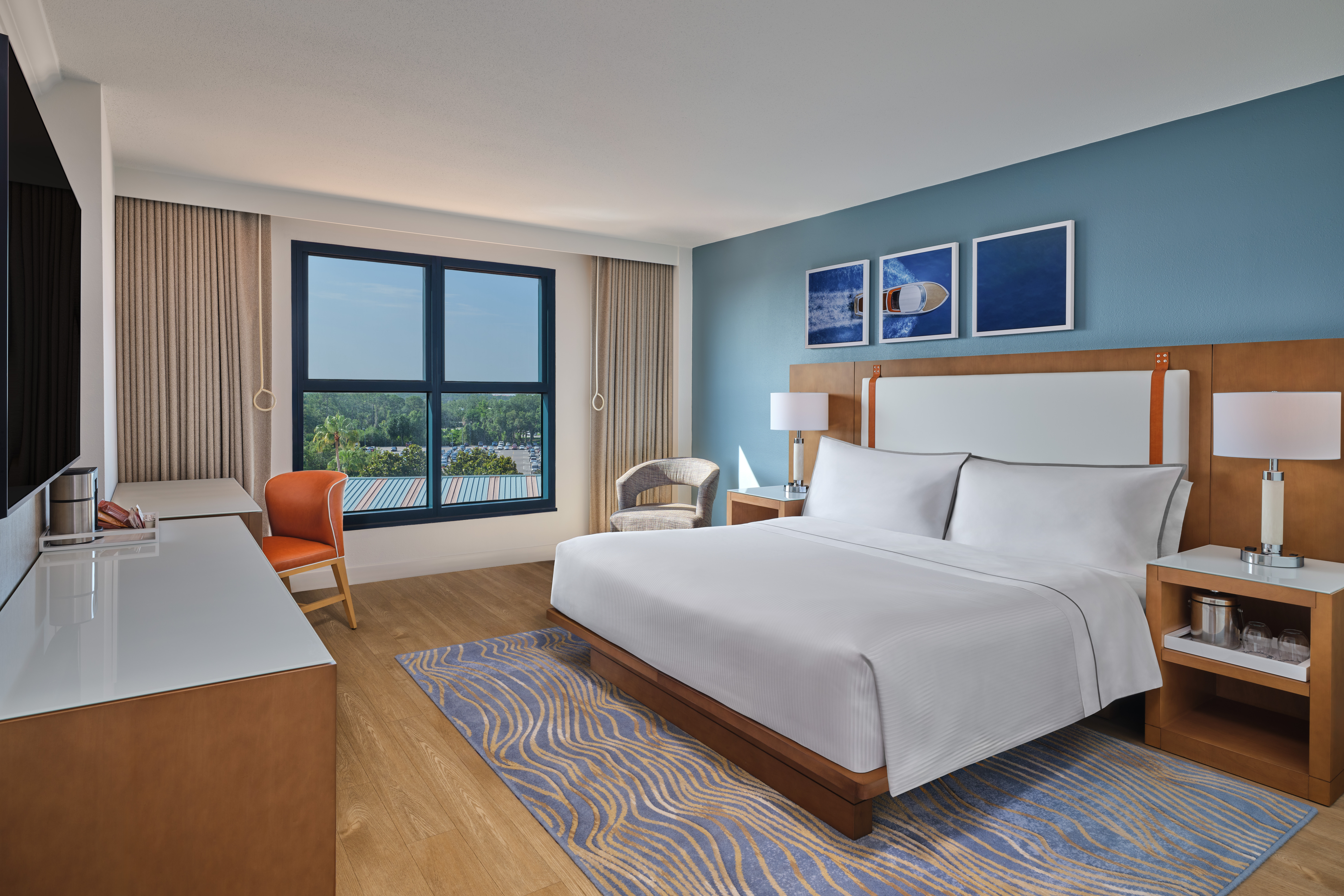 Modern hotel room with a large bed, blue accent wall, two nightstands, a TV, a desk with an orange chair, a wicker chair, and large window letting in natural light.