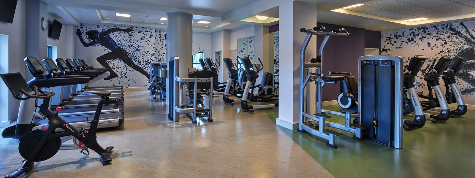 Modern gym with treadmills, stationary bikes, and weight machines. A running silhouette mural decorates the back wall, and the space is well-lit with natural and artificial light.