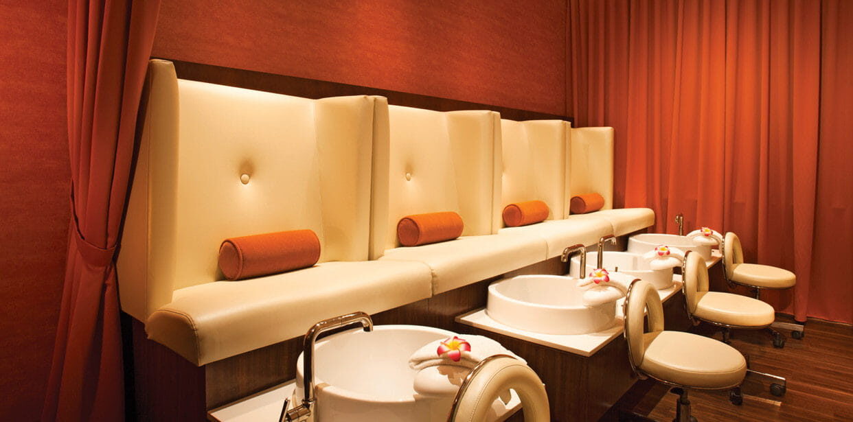 A spa room with four beige pedicure chairs, foot basins, and orange roll pillows, set against red walls and curtains.