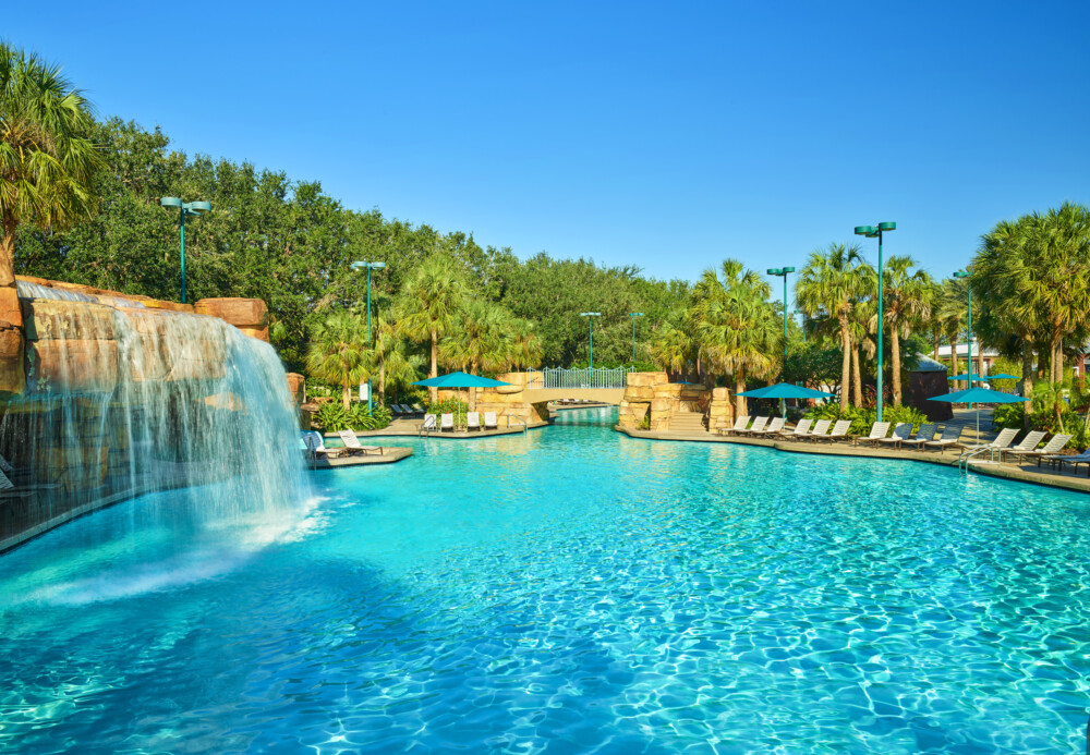 A large outdoor swimming pool with a waterfall, surrounded by palm trees, lounge chairs, and umbrellas under a clear blue sky.