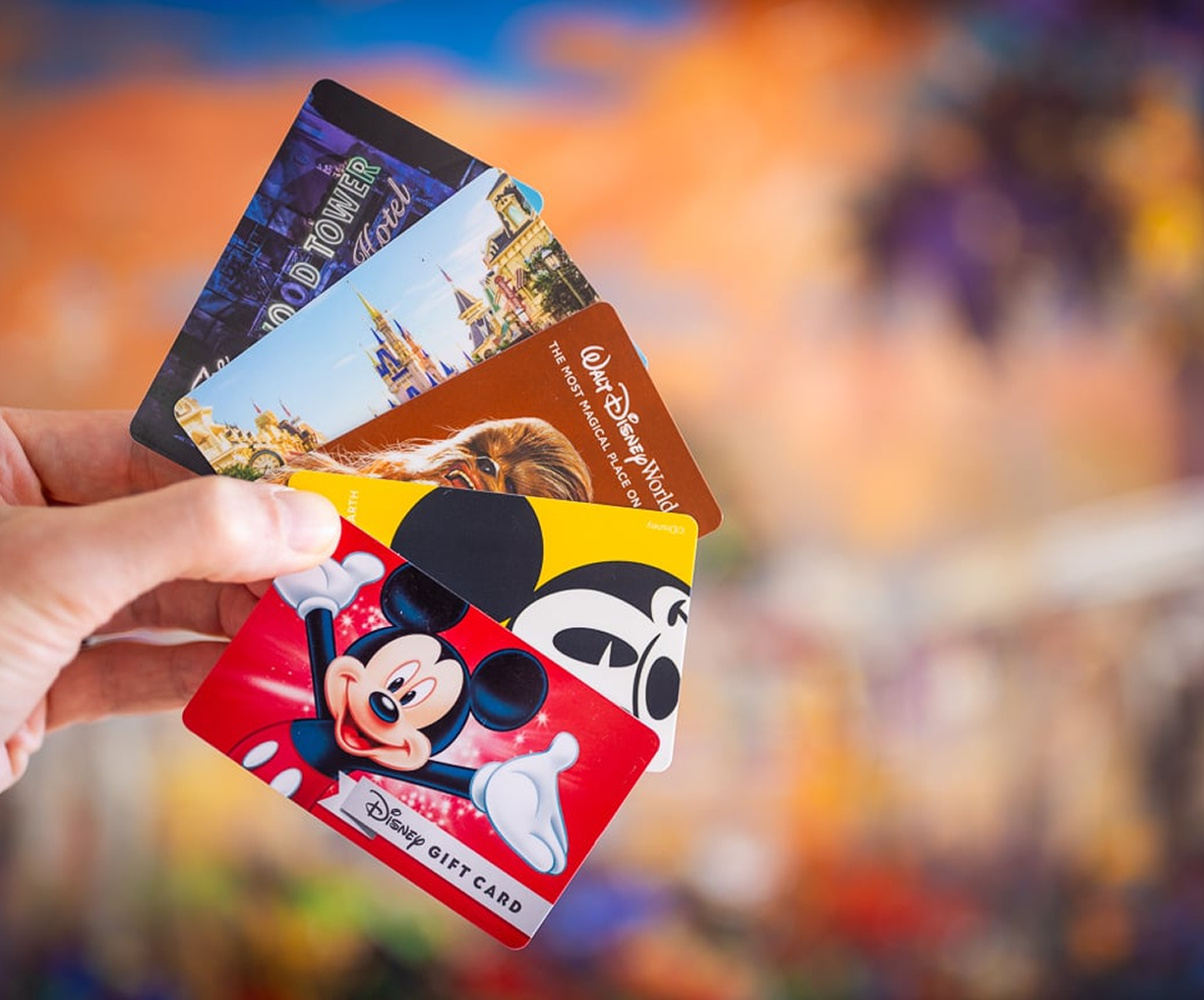 A hand holds four colorful Disney gift cards featuring Mickey Mouse, Chewbacca, Cinderellas Castle, and the Hollywood Tower Hotel with a vibrant, blurred background.