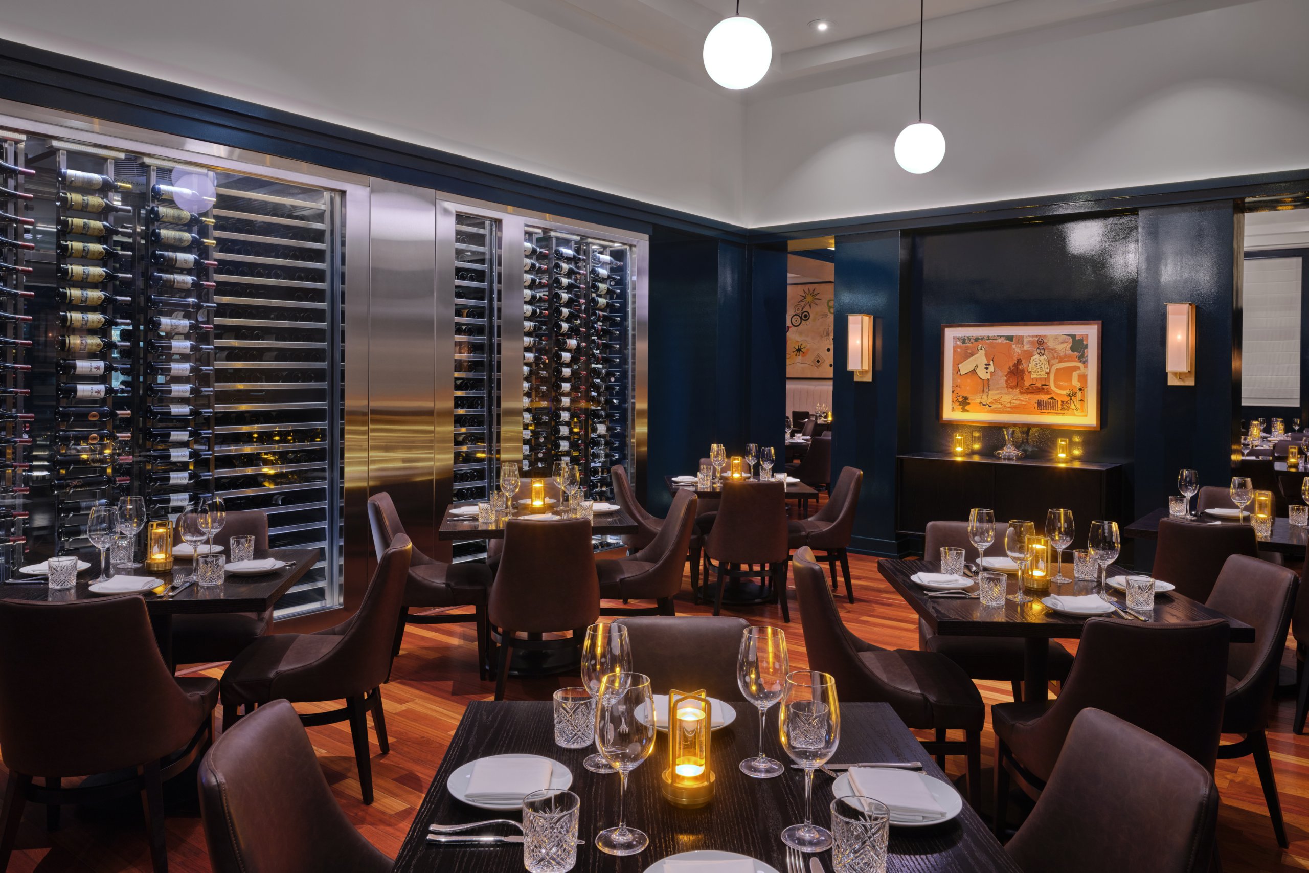 Elegant restaurant interior with dark wooden tables, brown chairs, soft candlelight, glassware, a modern wine rack on the left wall, and contemporary artwork. The atmosphere is warm and inviting with a sophisticated feel.