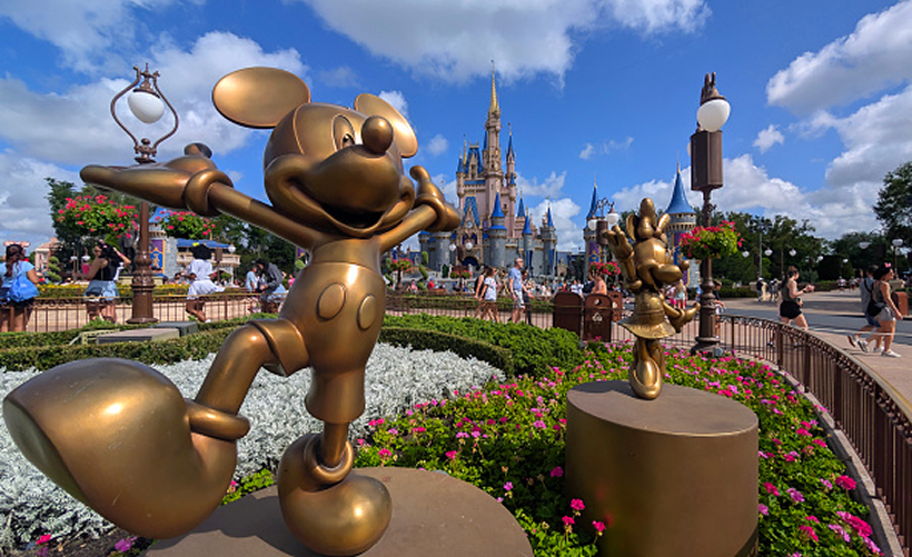 Bronze statues of cartoon mice stand in a colorful flower garden, with a castle and blue sky with clouds in the background. Visitors walk nearby, enjoying the sunny day at an amusement park.