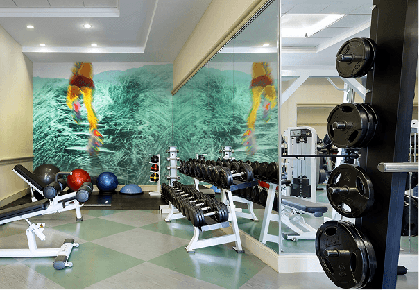 A modern gym with weights, benches, exercise balls, and mirrored walls. A vibrant mural of a person running on grass is featured on the back wall, adding energy to the workout space.