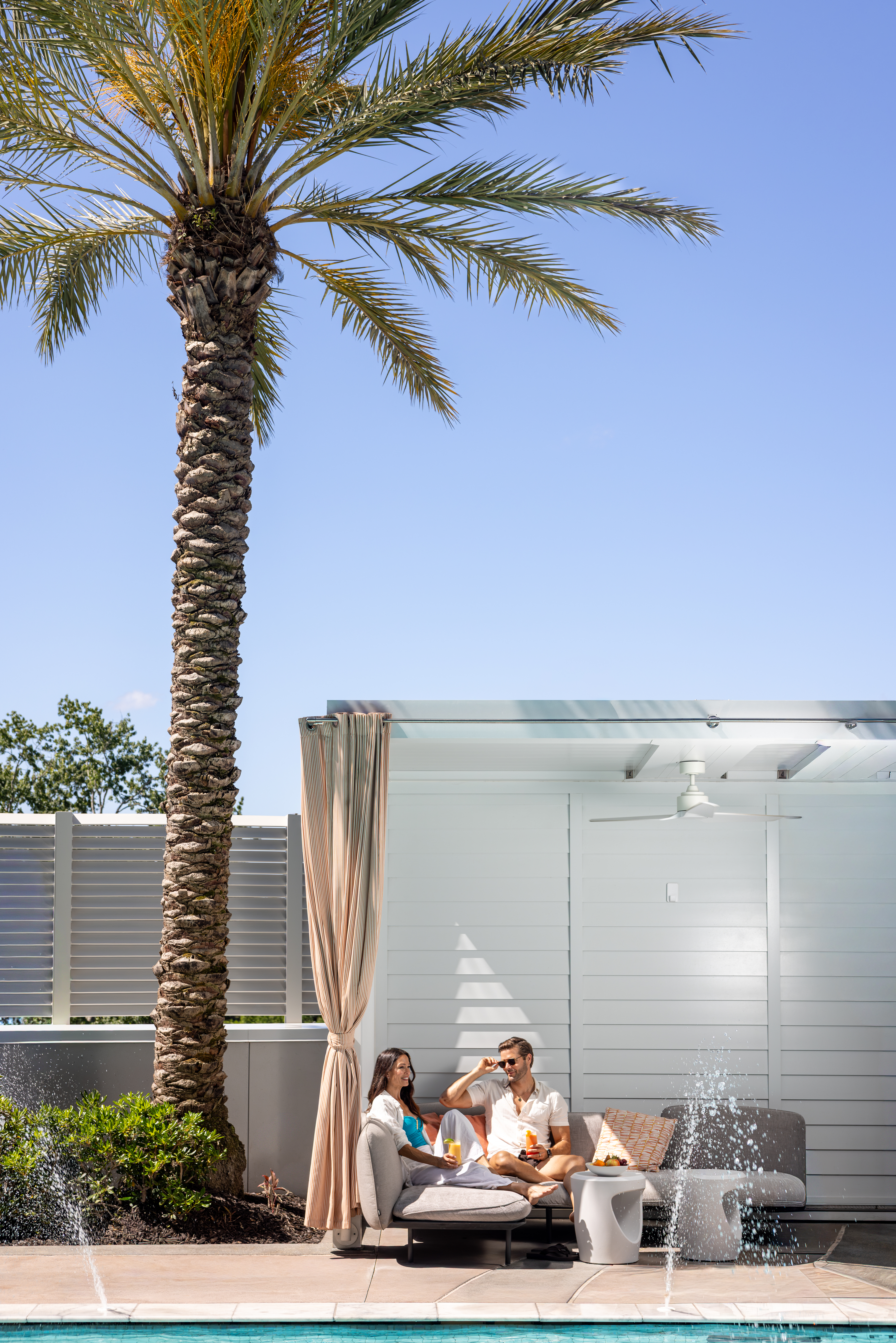 Couple sitting under a cabana with a palm tree on their left and water springing from the pool.