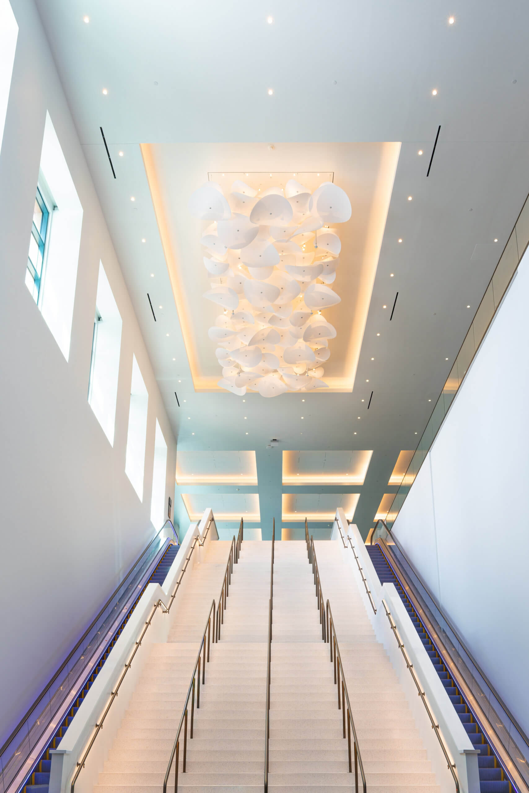 A wide, well-lit staircase with glass railings ascends under modern white ceiling lights and large windows on the left, creating a bright, spacious interior with clean architectural lines.