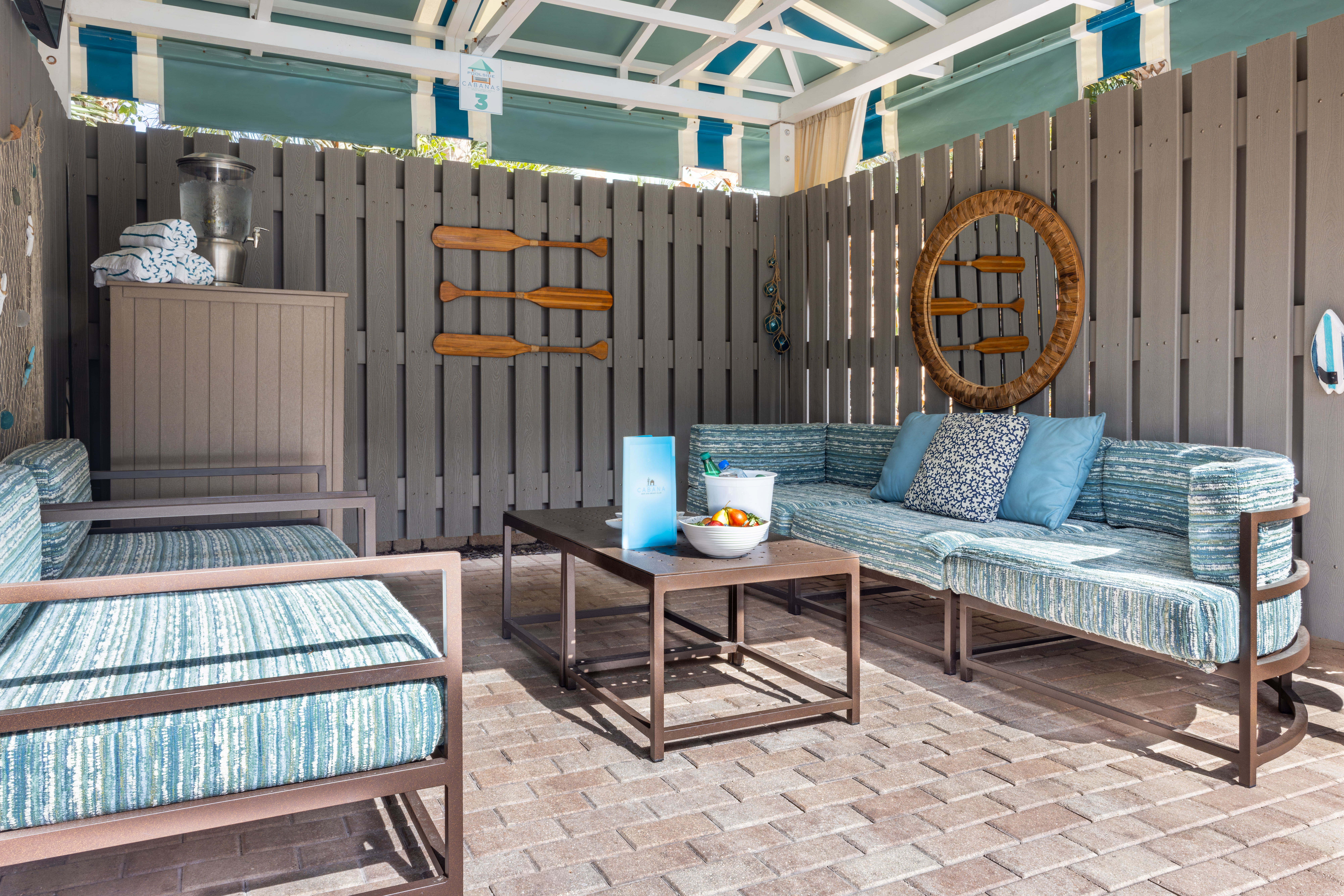 A cabana with a blue couch and two chairs, table, towels and fruit and water on the table.