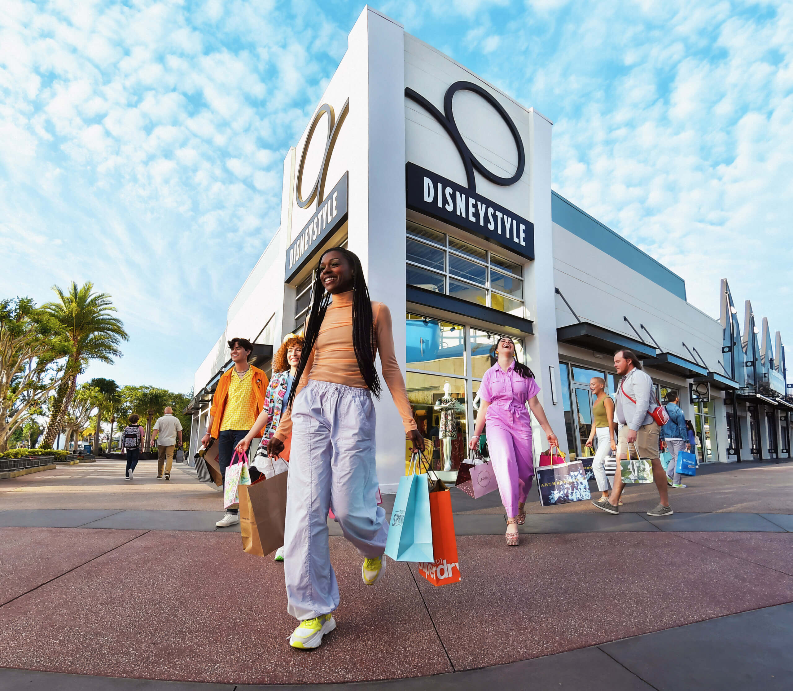 Smiling people exit a DisneyStyle store with shopping bags under a bright blue sky. Palm trees line the sidewalk, and the building features large DisneyStyle signs.