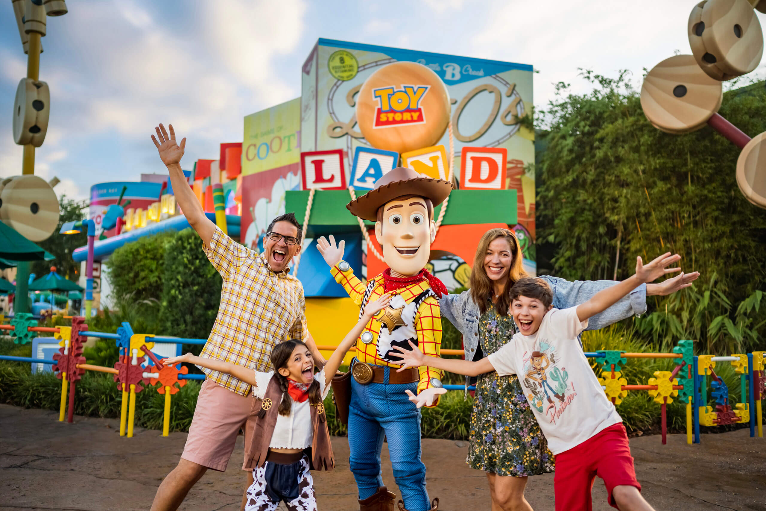 A family of four poses excitedly with Woody, the cowboy from Toy Story, at Toy Story Land. Bright, colorful decorations and signs fill the background, creating a playful and cheerful atmosphere.