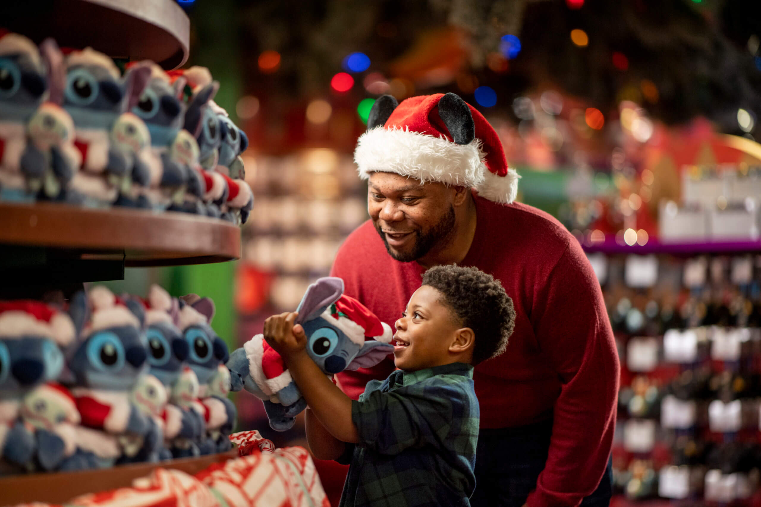 A man in a Santa hat and a young boy smile while looking at Christmas-themed stuffed toys in a store. The child holds up one of the plush toys excitedly.