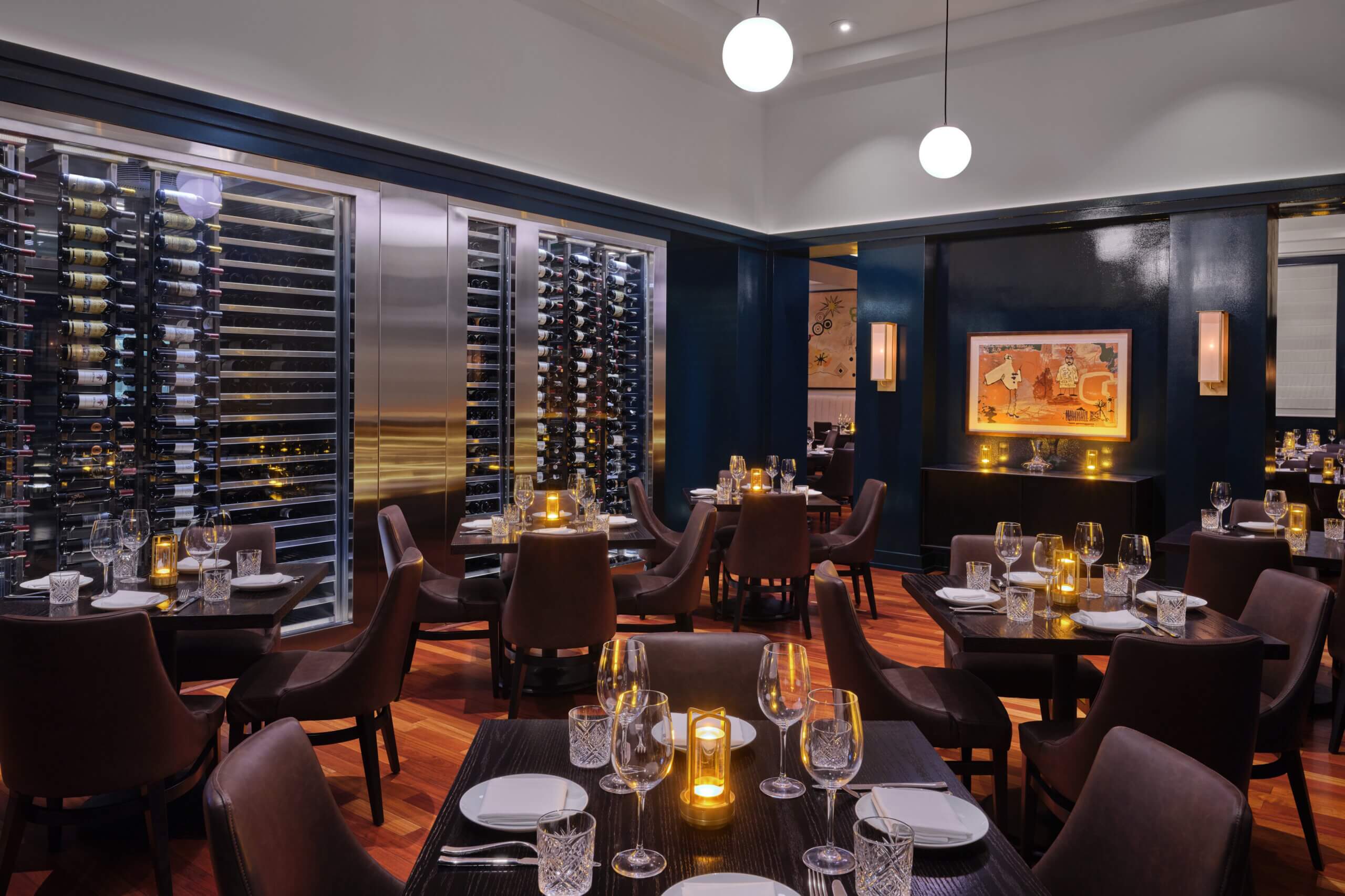 Elegant restaurant interior with dark wood tables, cushioned chairs, wine racks, modern lighting, and softly glowing candles. Artwork and wine display add to the upscale ambiance.