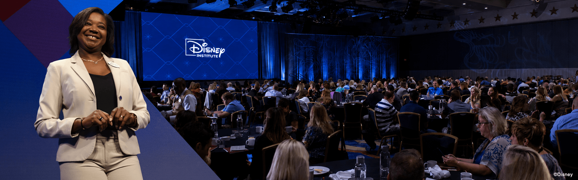 A woman in a suit stands smiling on stage left; in the background, a large audience sits at tables facing a screen displaying the Disney Institute logo inside a conference room.