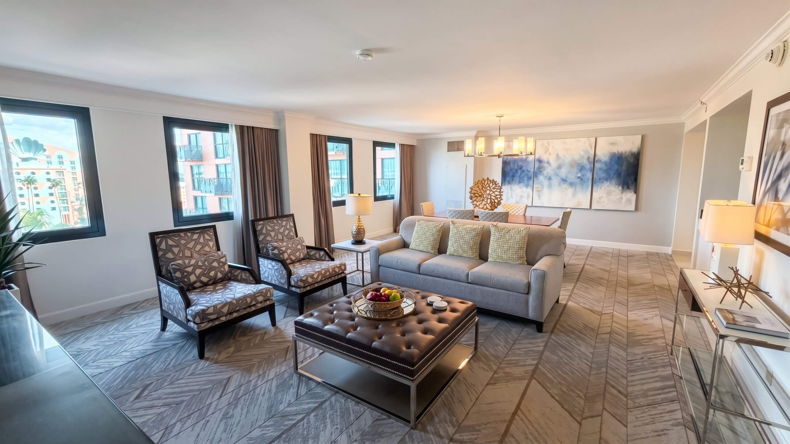 Modern living room with large windows, a gray sofa, two patterned armchairs, a brown ottoman with fruit, lamps, and a dining area in the background with contemporary art on the wall.
