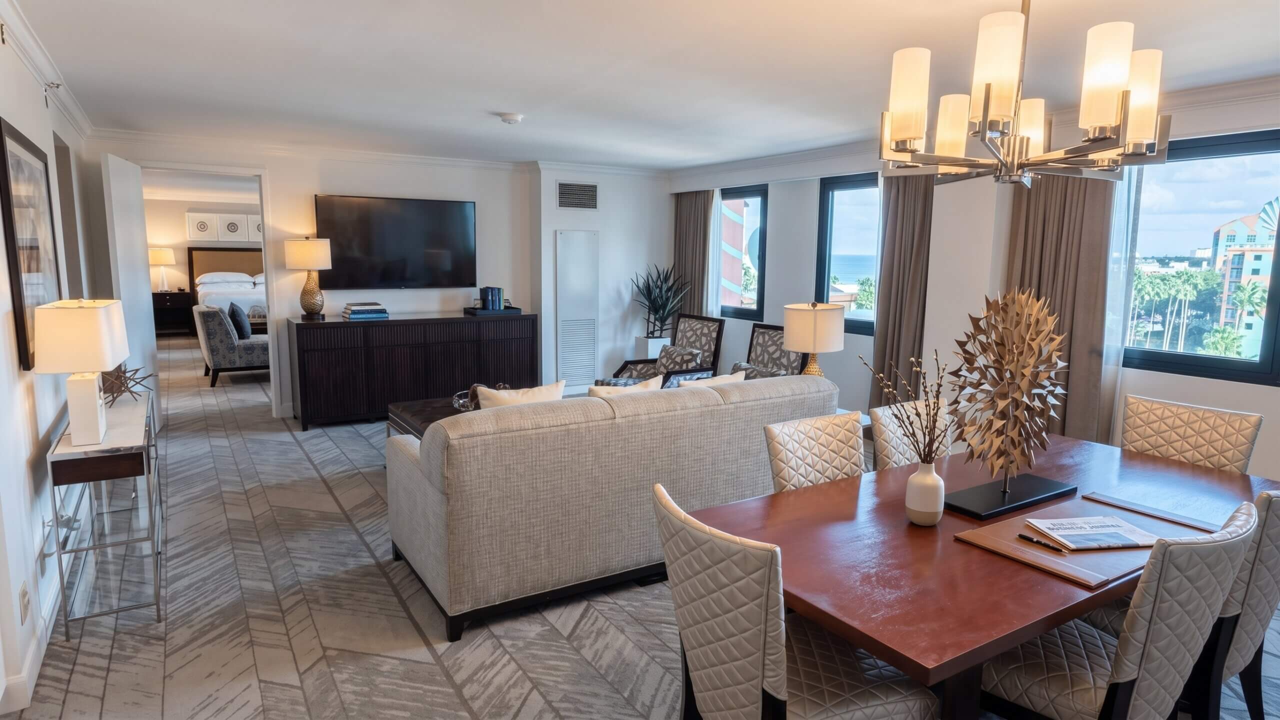 Modern hotel suite with a living area, TV, bedroom in the background, dining table with chairs, large windows, and neutral-toned decor. Natural light fills the space, highlighting elegant furnishings and contemporary design.