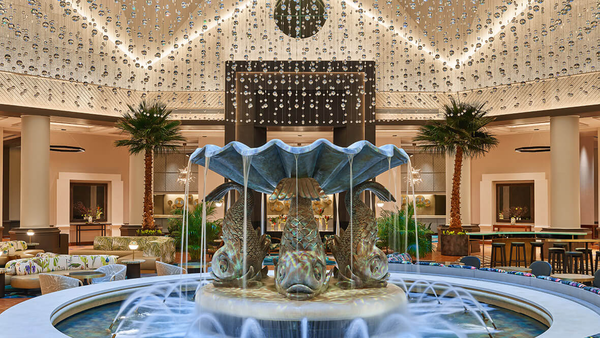 Luxurious hotel lobby with a large sculpted fountain featuring fish, surrounded by palm trees, seating areas, and hanging crystal decor from the ceiling.