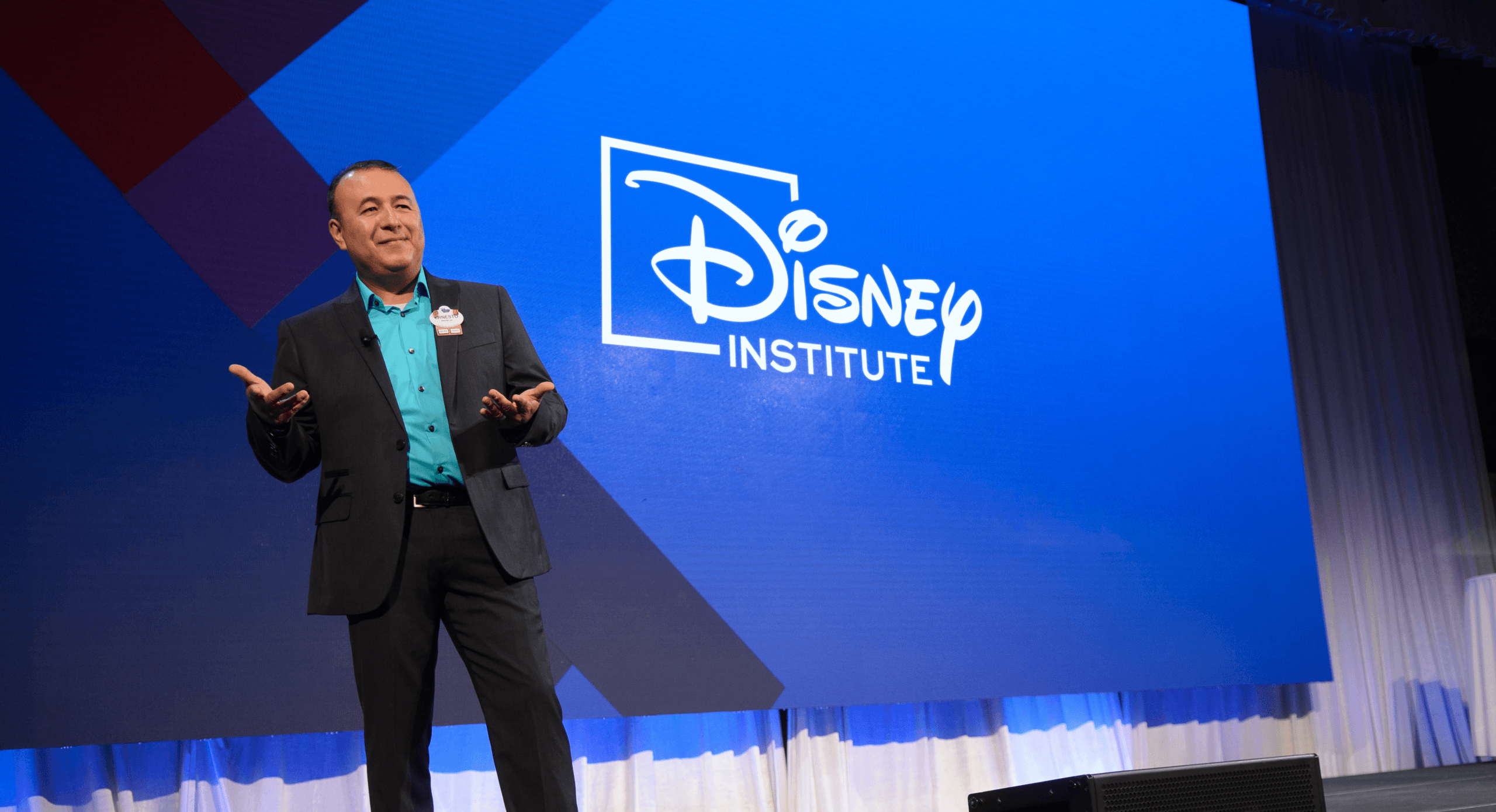 A man in business attire stands on stage, gesturing with his hands, in front of a large screen displaying the Disney Institute logo.