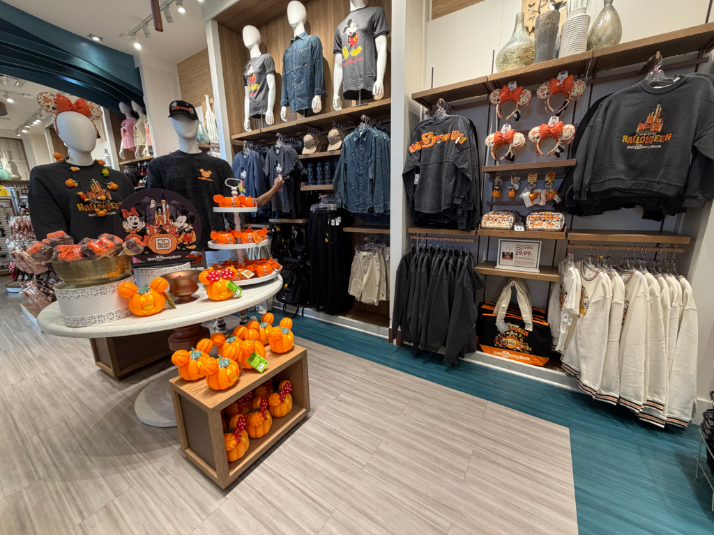 Une vitrine de magasin présentant des articles Mickey Mouse sur le thème d'Halloween, notamment des t-shirts, des pulls, des mugs, des citrouilles en peluche et des friandises. La décoration comprend des mini-citrouilles orange et des éléments automnaux disposés sur les étagères et les tables.