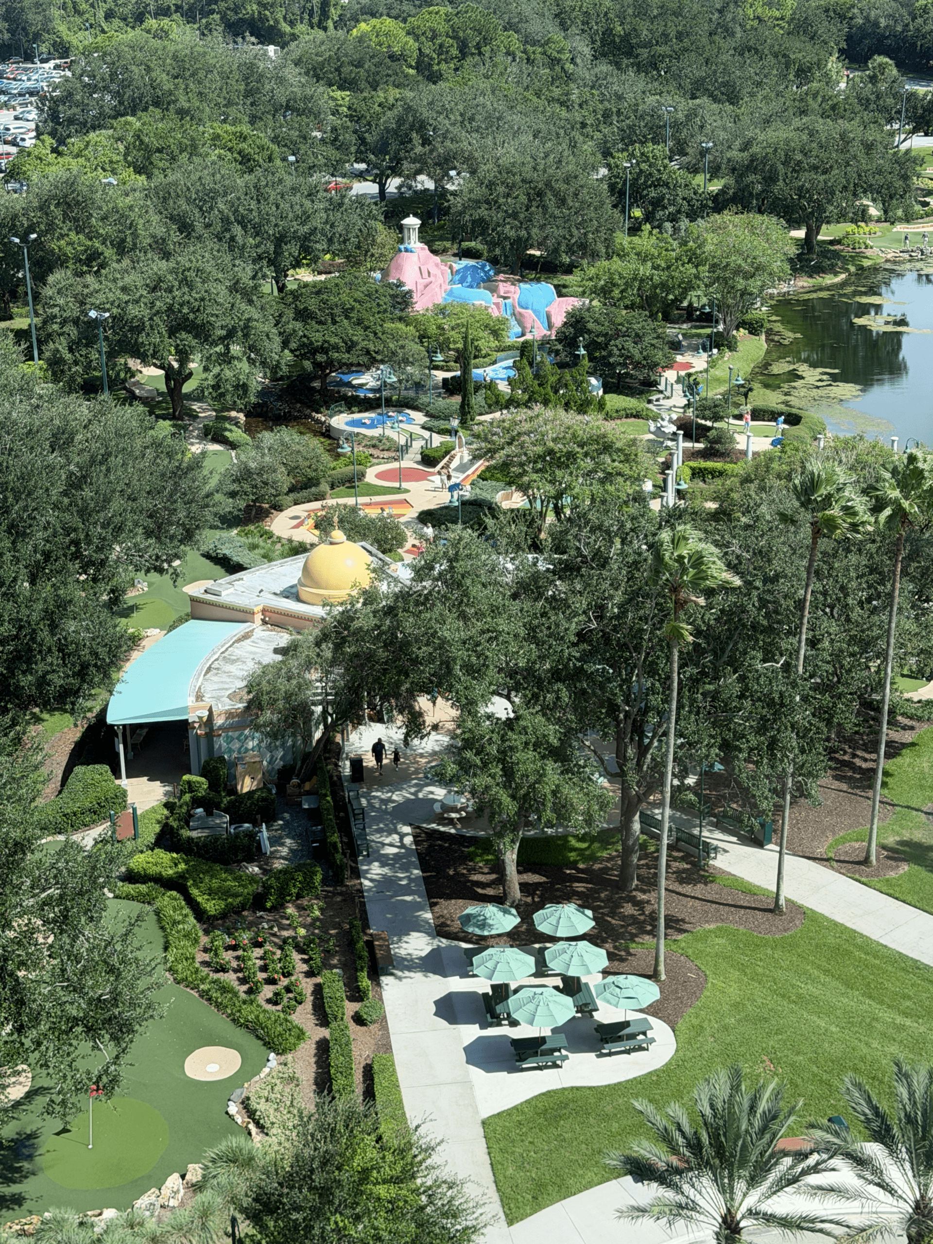 Vista aérea de un campo de minigolf con sombrillas verdes, árboles, senderos, un estanque y coloridas estructuras de juegos acuáticos al fondo. La zona recreativa está rodeada por un aparcamiento y un bosque frondoso.
