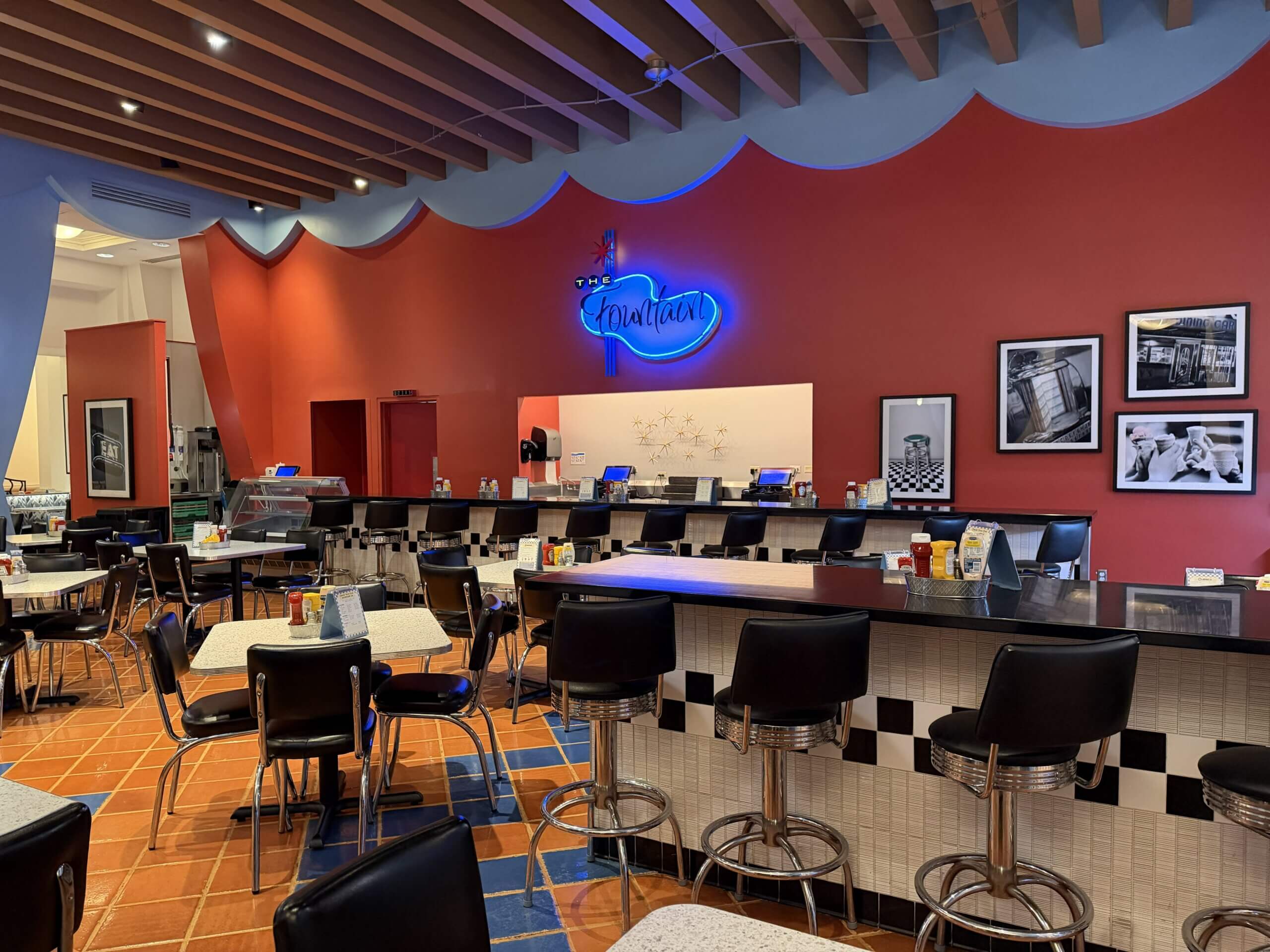 Un restaurant rétro avec des tabourets de bar noirs, des comptoirs à carreaux, des murs rouges et des photos en noir et blanc. Une enseigne au néon bleue indique « Chantarelle ». Les tables et le bar sont vides.