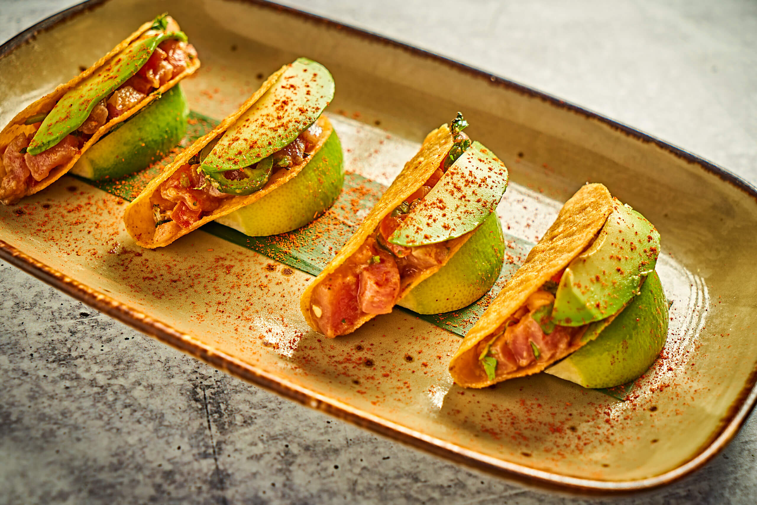 Three mini tacos filled with diced meat and avocado slices are arranged upright on lime halves, served on a rectangular plate with a sprinkle of red spice.