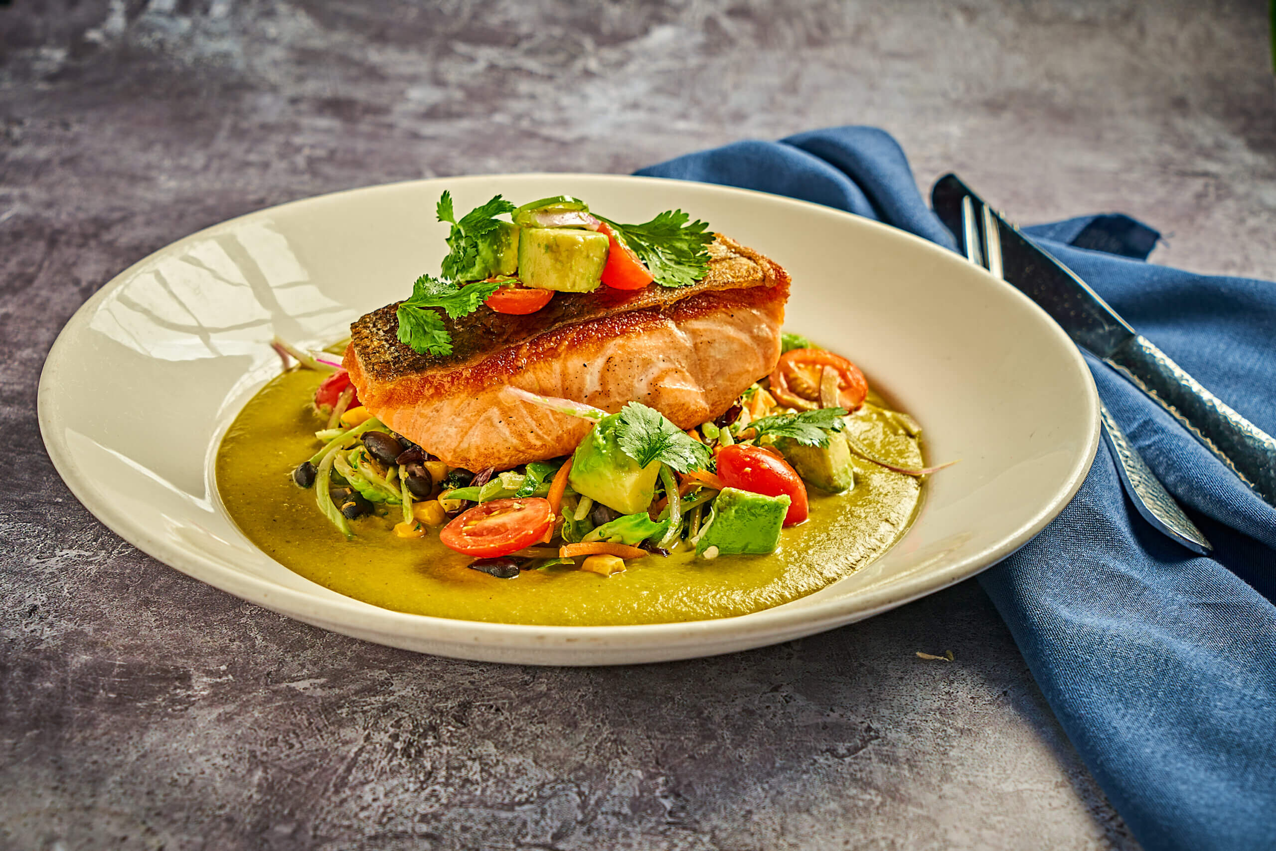 A plate with seared salmon on a bed of vegetables, avocado, and cherry tomatoes, garnished with cilantro, served over a green sauce. A fork and knife rest on a blue napkin beside the plate.