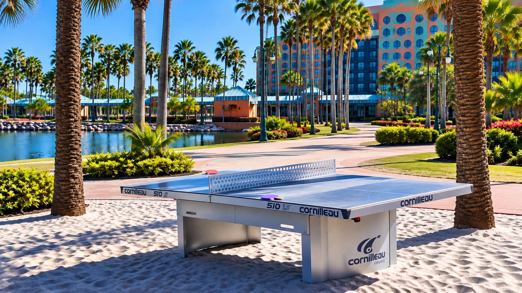 A ping pong table sits on sand among palm trees near a lake, with a colorful building and blue sky in the background, creating a tropical resort atmosphere.