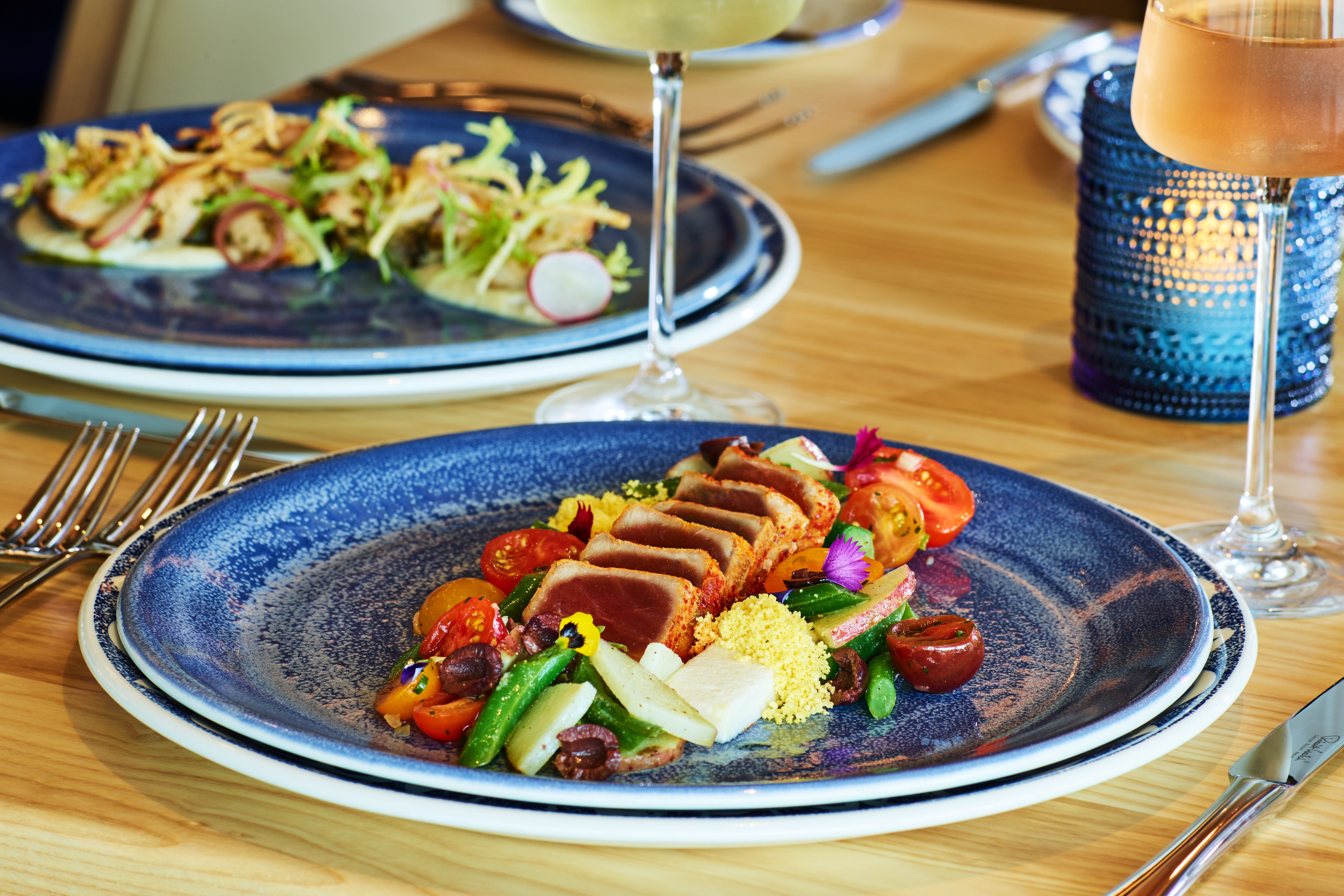 A blue plate with seared tuna, cherry tomatoes, asparagus, olives, and garnishes sits on a wooden table with a salad, wine glasses, and a decorative blue candle holder in the background.