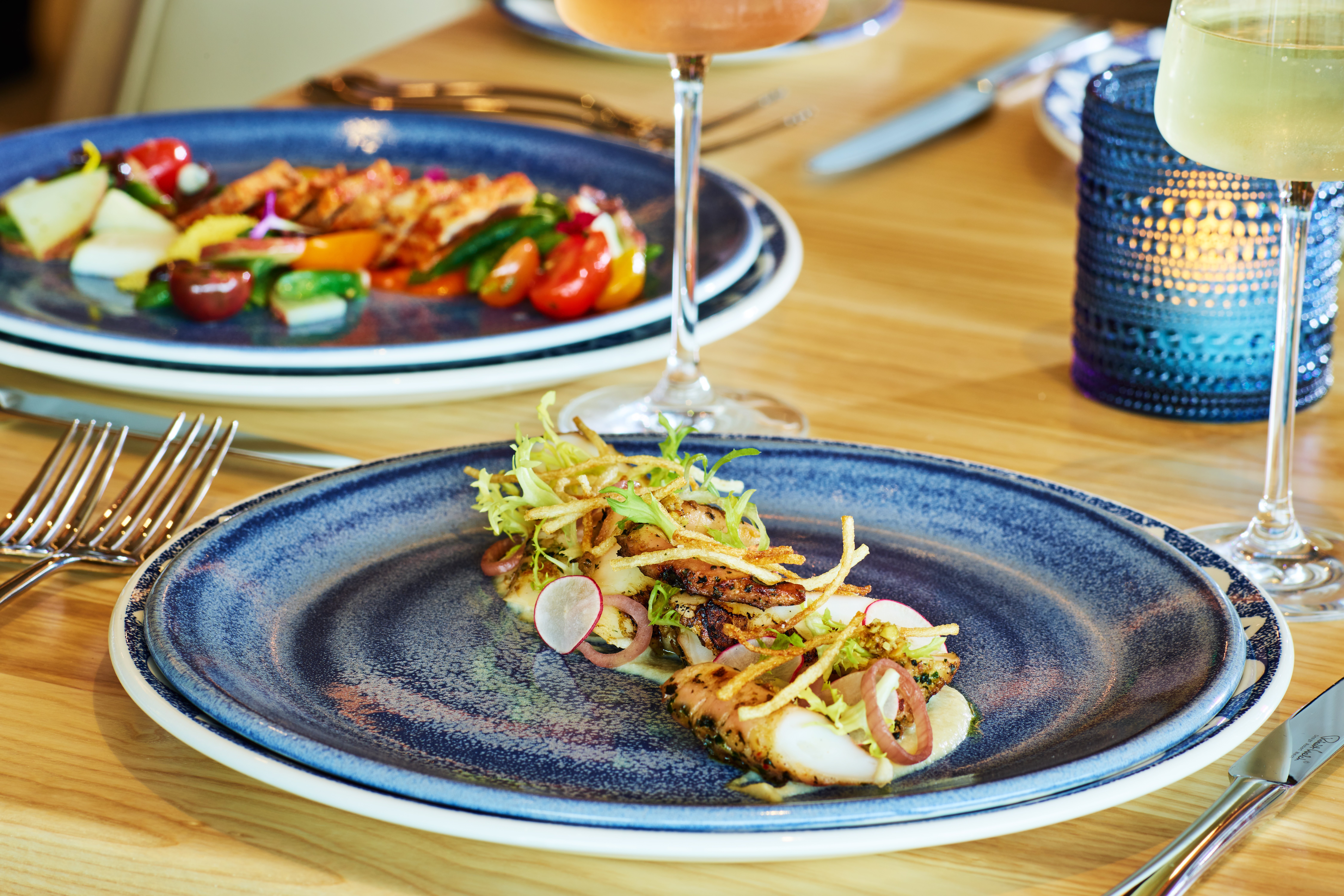 A gourmet dish with grilled fish, crispy garnishes, and radish slices on a blue plate, with a colorful salad and two wine glasses in the background on a wooden table.
