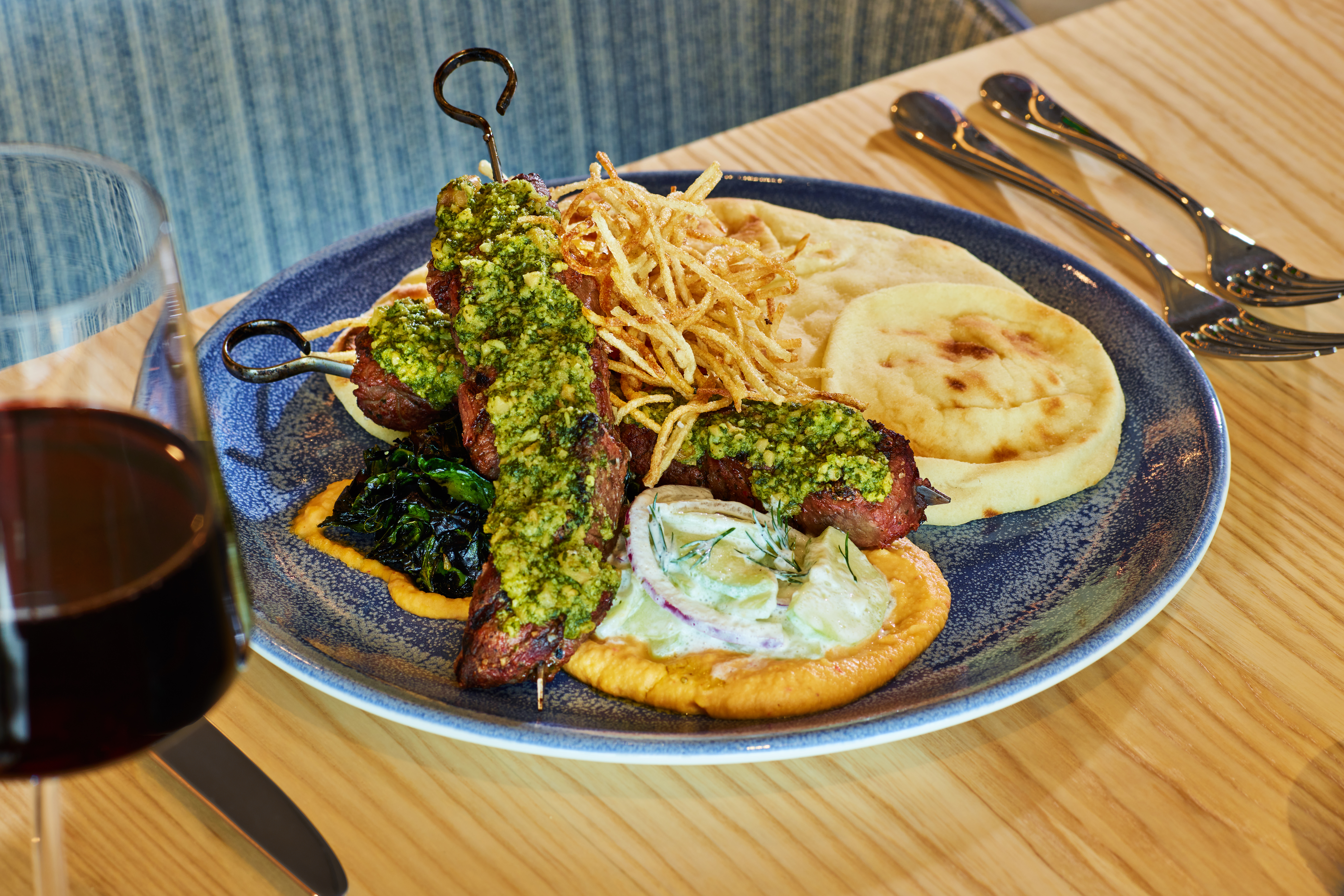 Two meat skewers with green sauce on a blue plate, served with thin fries, pita bread, creamy sauce, greens, and a glass of red wine on a wooden table with cutlery nearby.