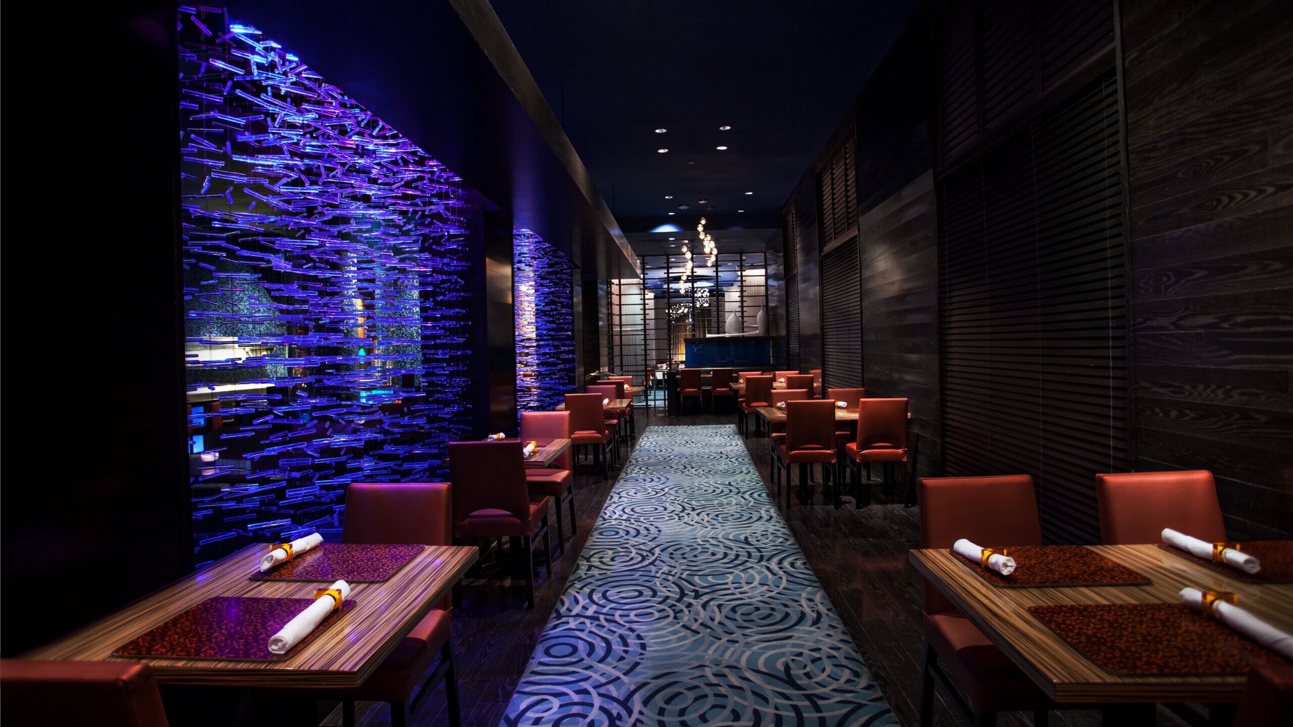 Elegant restaurant interior with red chairs, wooden tables set with napkins, a blue illuminated wine rack on one wall, patterned carpet, and dim, modern lighting. The atmosphere is stylish and contemporary.