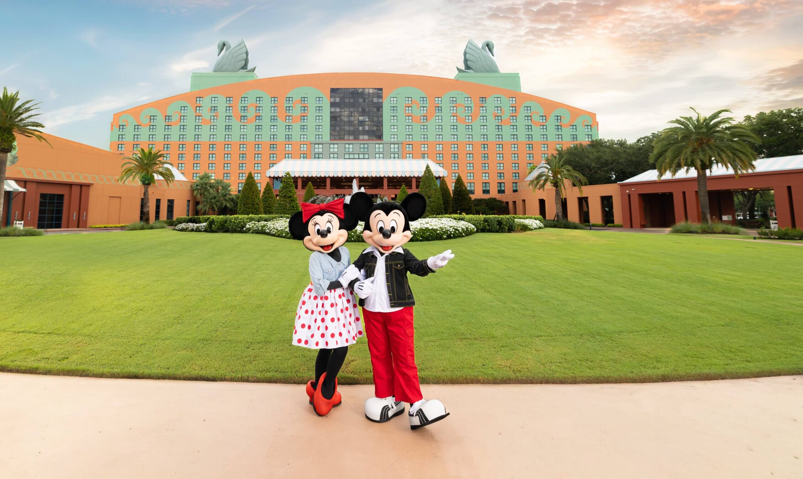 Minnie Mouse and Mickey Mouse stand together on a sidewalk, posing in front of a large, colorful hotel with distinctive swan statues on the roof and a well-kept lawn under a partly cloudy sky.