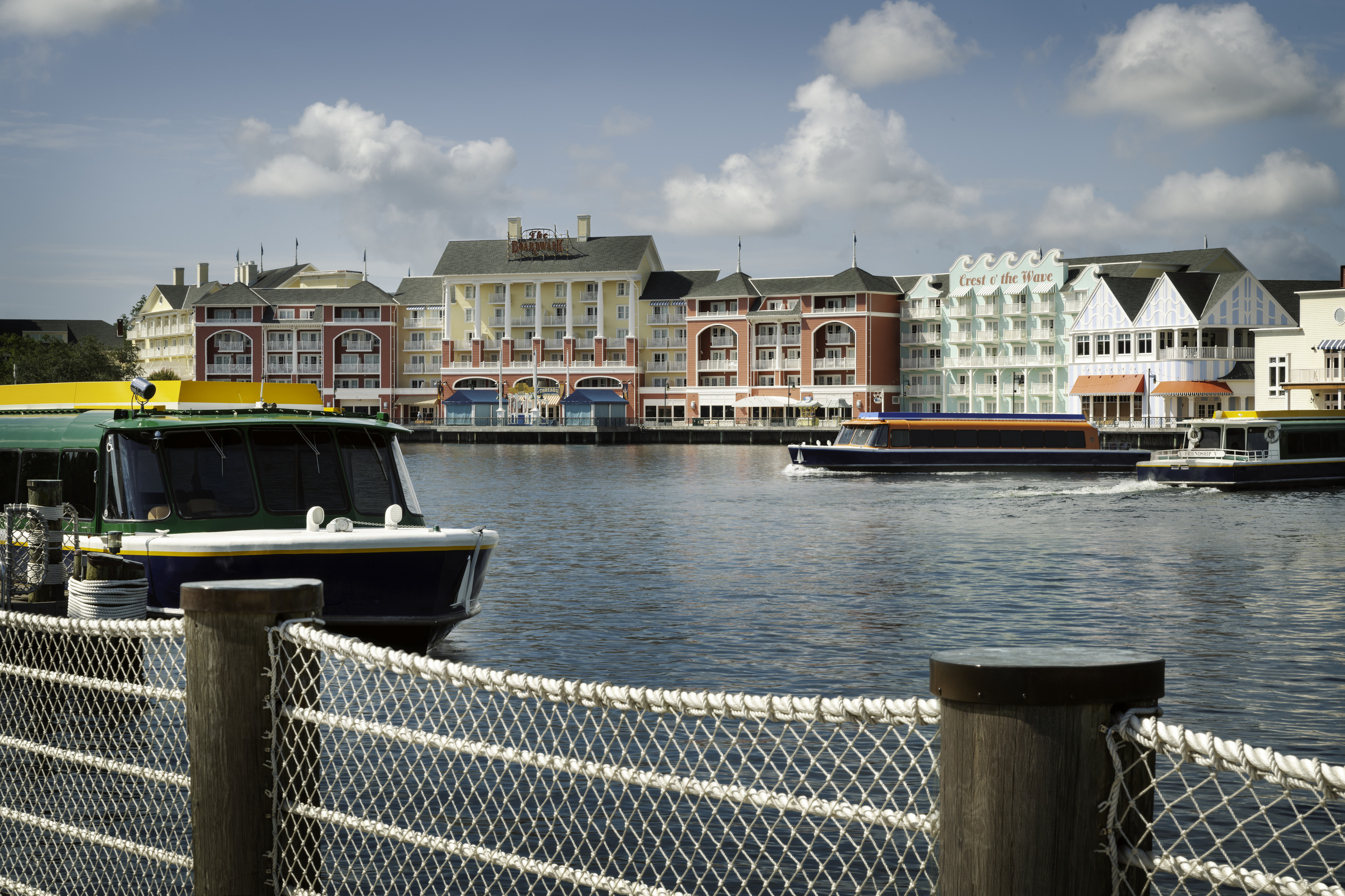 A picture of several buildings painted yellow, peach and light green, with three Disney boats in front of it on the lake.