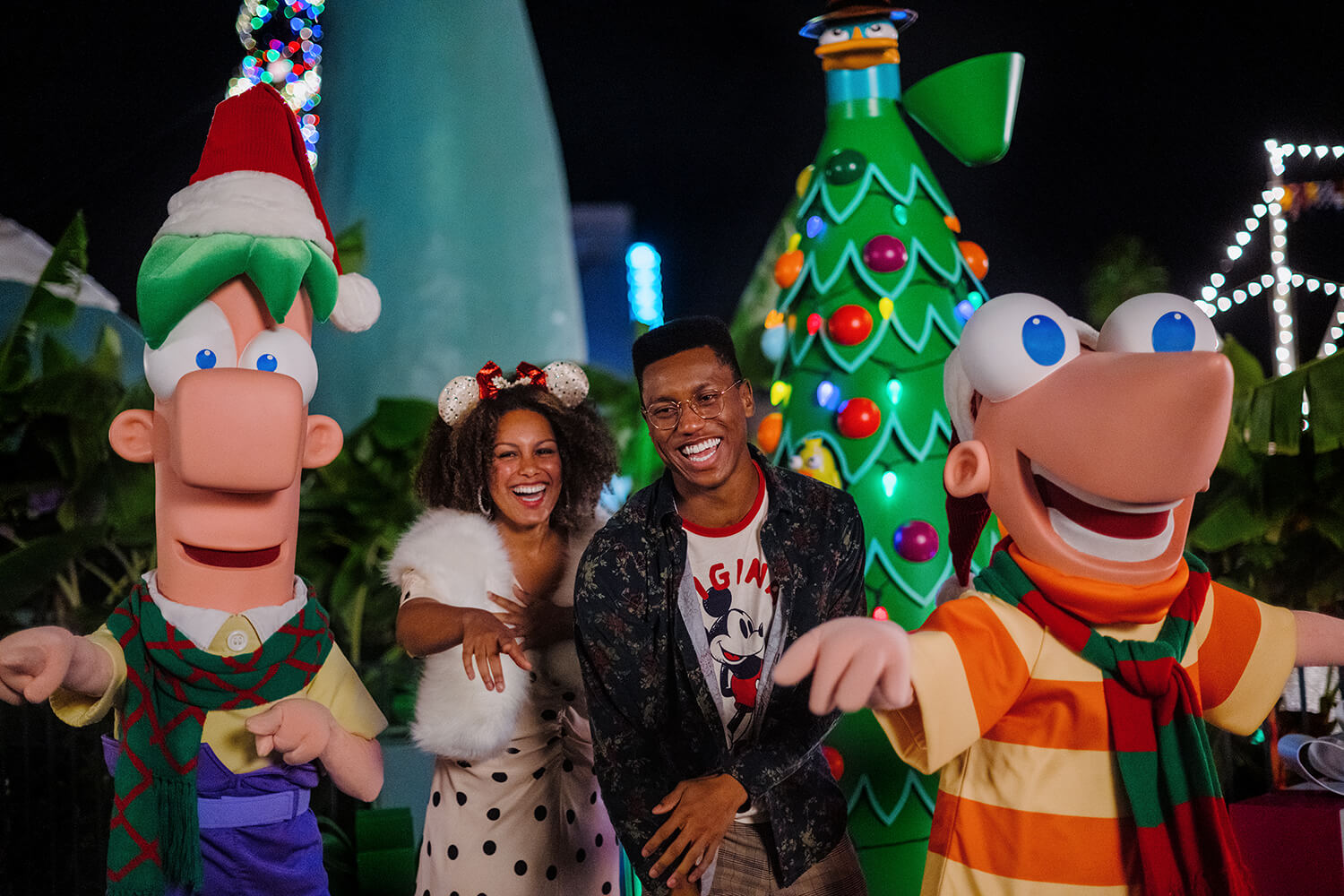 Deux personnes sourient et prennent la pose aux côtés de deux mascottes représentant des personnages de dessins animés, vêtues de tenues de fête, devant des décorations de Noël, notamment un grand sapin et des guirlandes lumineuses qui scintillent dans la nuit.