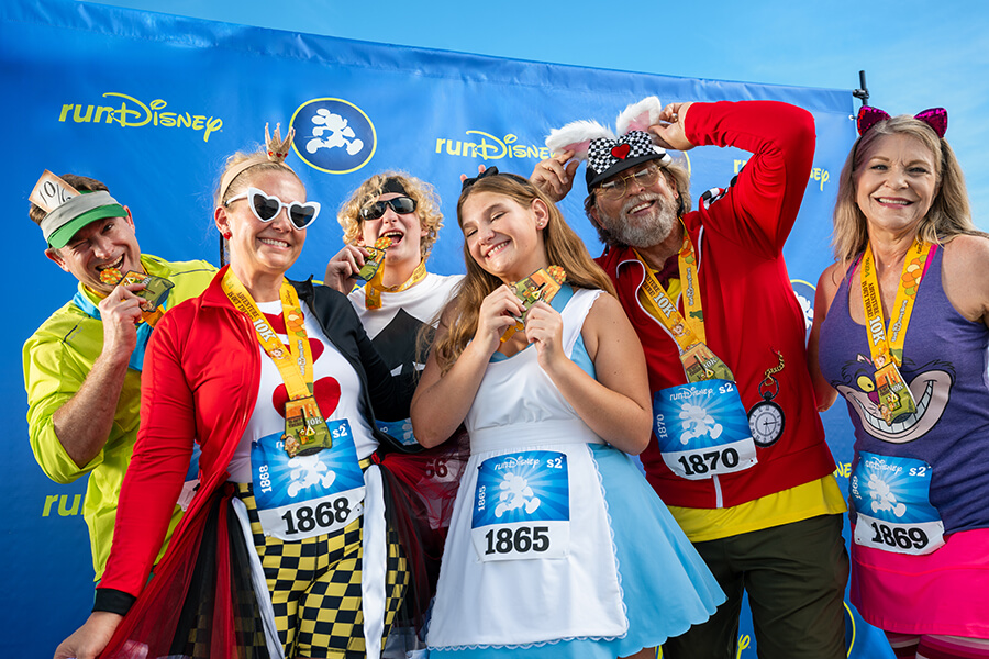 Seis adultos sorridentes, vestidos com fantasias coloridas de “Alice no País das Maravilhas”, exibem orgulhosamente suas medalhas em frente a um cenário azul da runDisney após concluírem uma corrida.