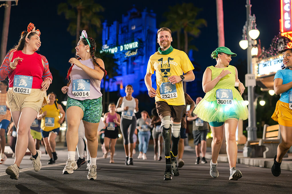 A group of happy runners in colorful costumes, including animal ears, a tutu, and a prosthetic leg, run at night past the Hollywood Tower Hotel sign. Other participants are in the background.