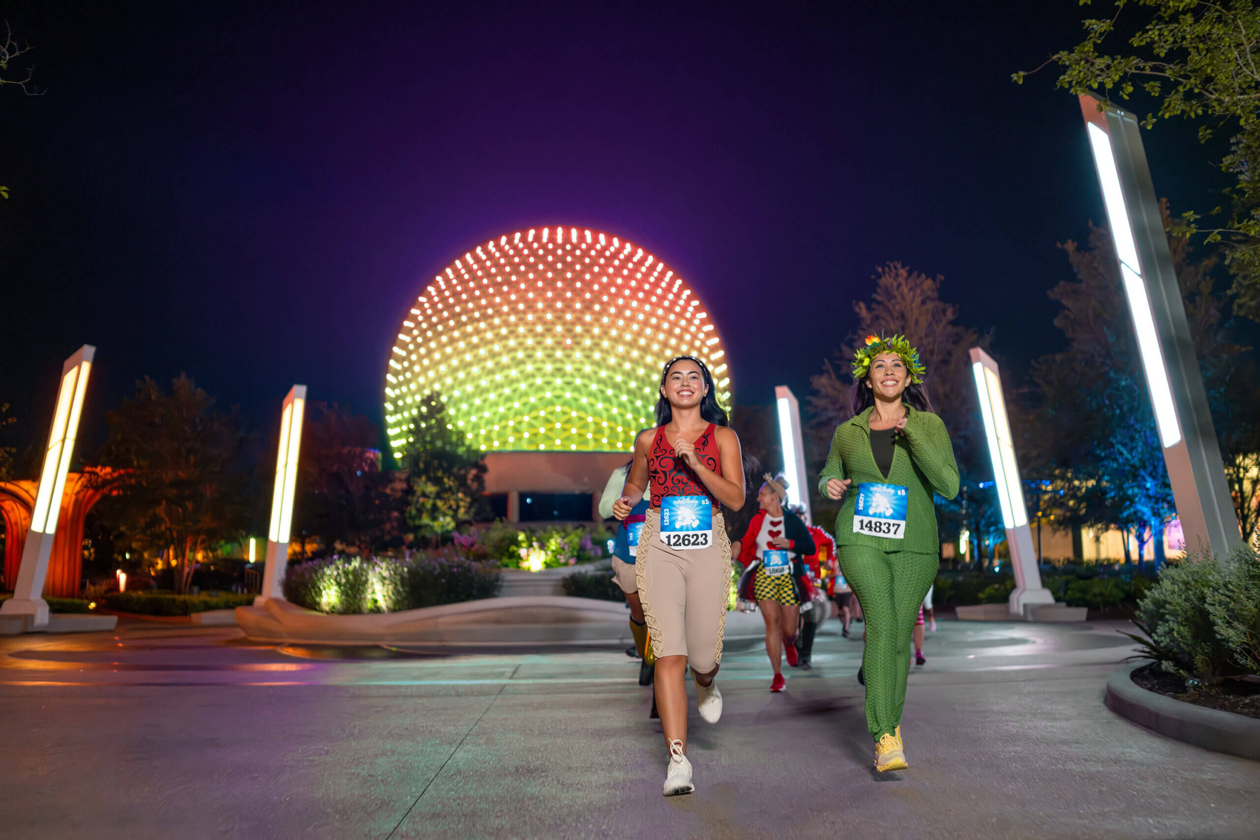 Two women in costumes run at night in a race, smiling, with a glowing, spherical building and colorful lights in the background. Other runners follow behind them.