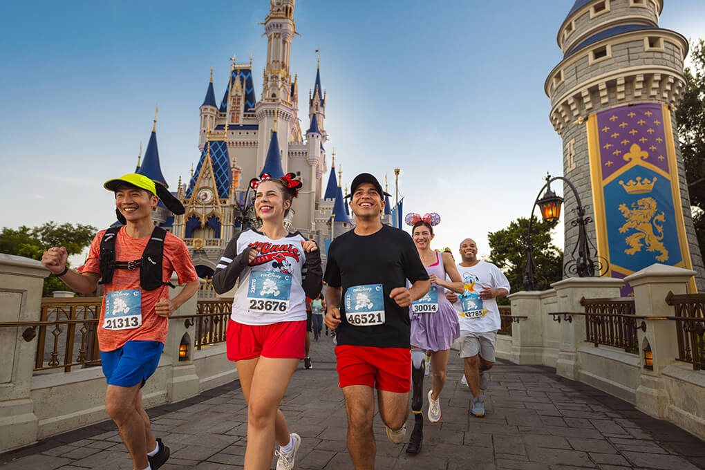 Five people in running gear, some wearing Disney-themed outfits and bibs, smile as they run past a castle at a theme park on a sunny day.