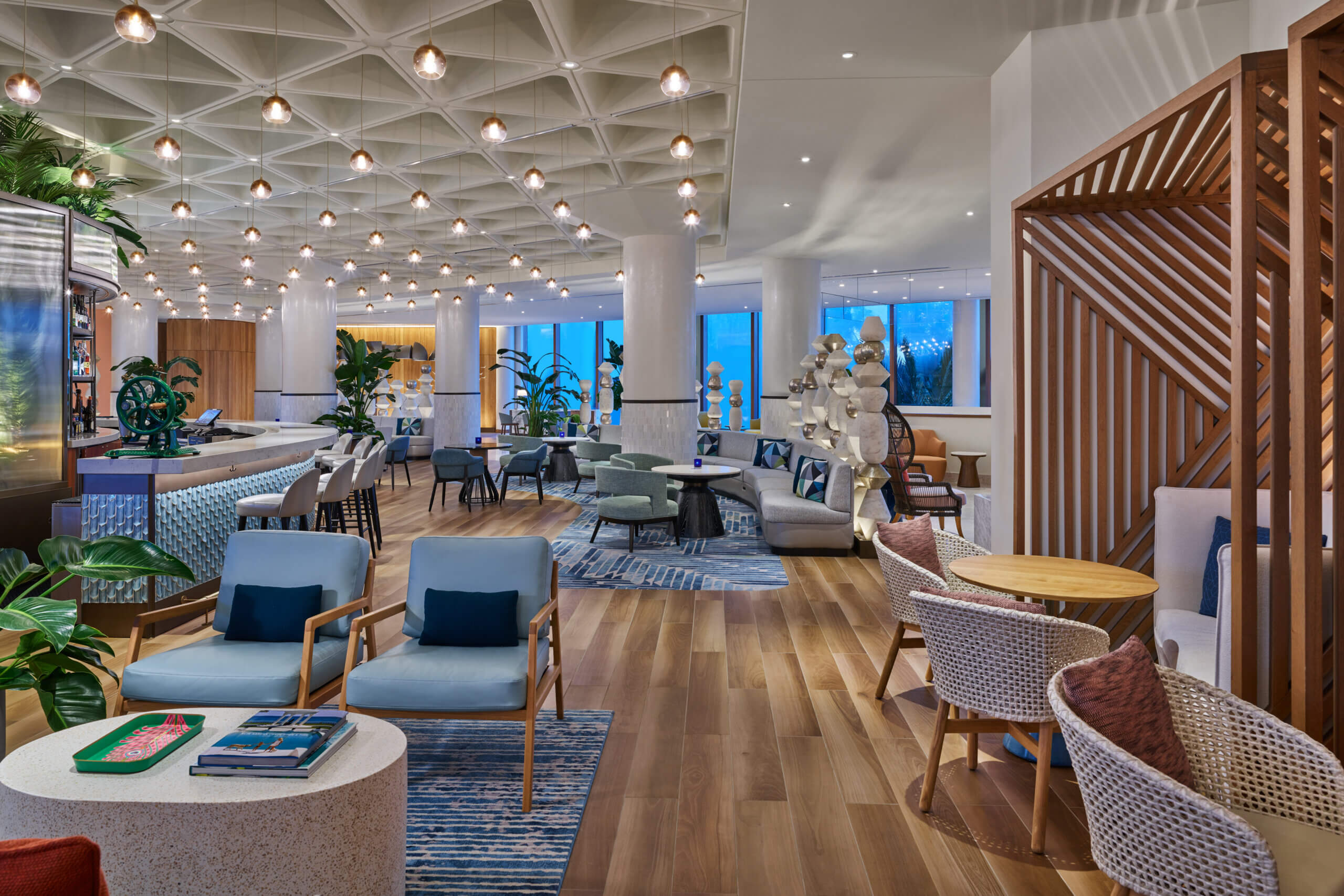 Modern hotel lounge with cozy seating, wood floors, blue accents, plants, pendant lights, and large windows letting in natural light. Stylish décor and a relaxed, inviting atmosphere.