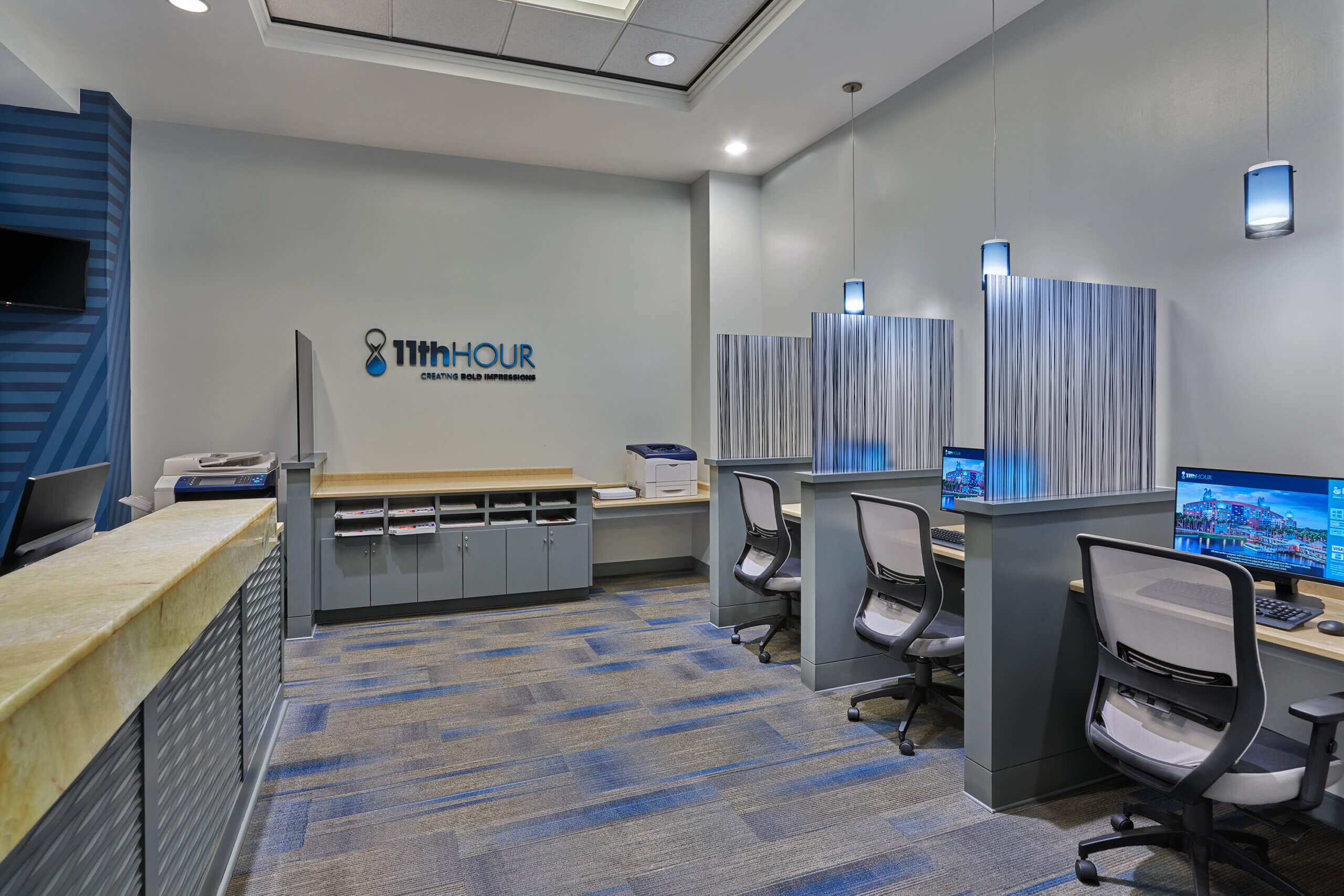 Espacio de oficinas moderno con puestos de trabajo informatizados separados por mamparas, sillas ergonómicas, lámparas colgantes y un mostrador con armarios de almacenamiento frente a una pared de color gris claro.