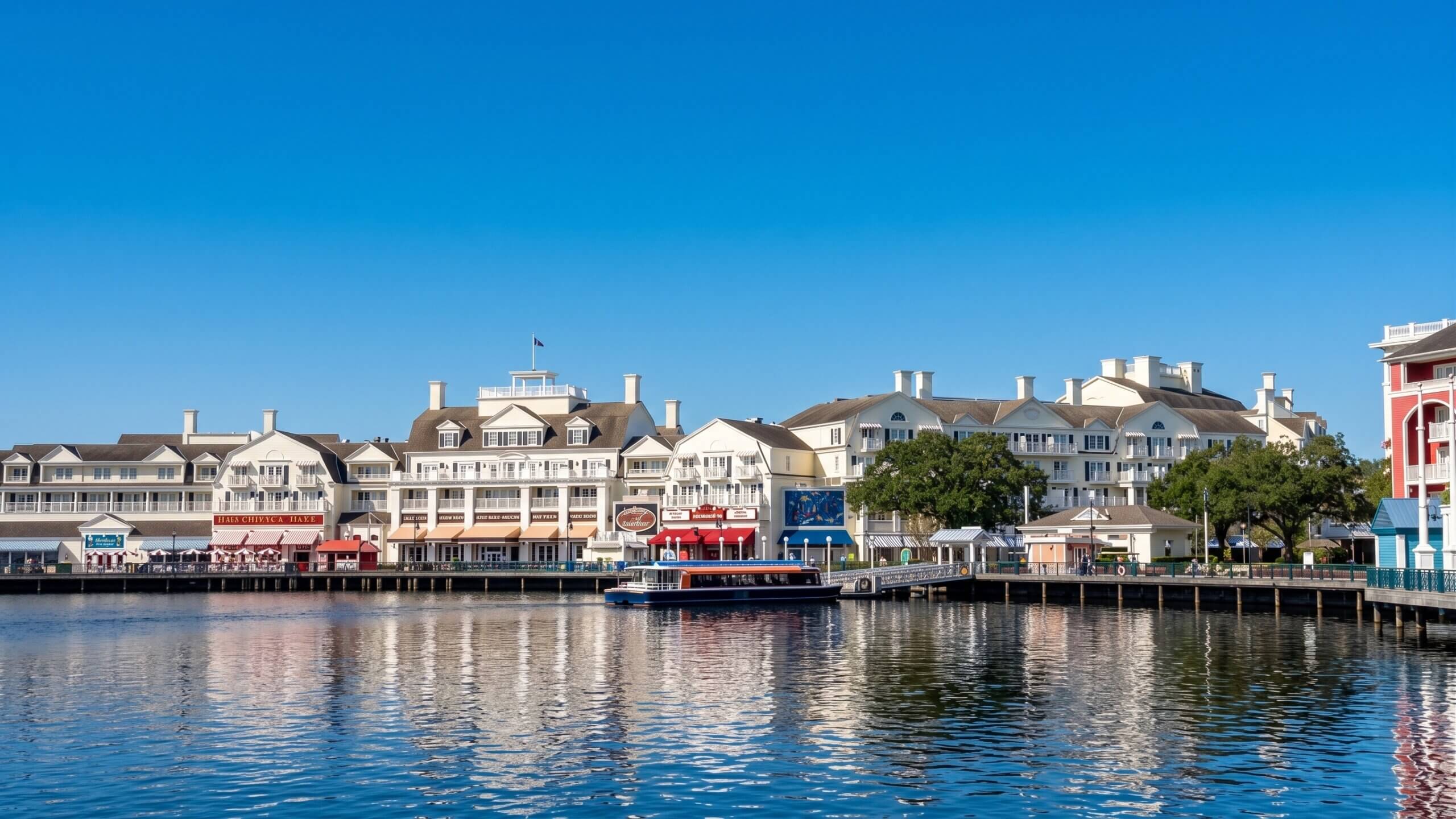 A waterfront scene with white, resort-style buildings, restaurants, and shops reflected in calm water under a bright blue sky. A boat is docked along the wooden boardwalk.