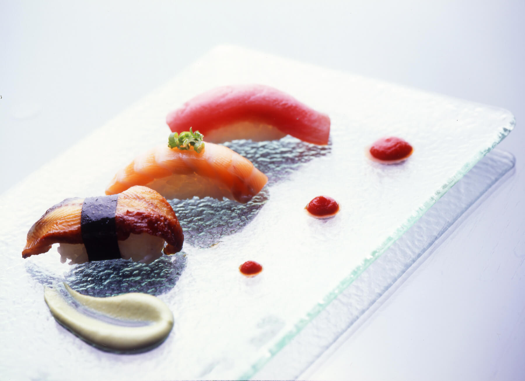 Three pieces of sushi on a rectangular glass plate, garnished with small dollops of red sauce and a swirl of green sauce, set on a light background.