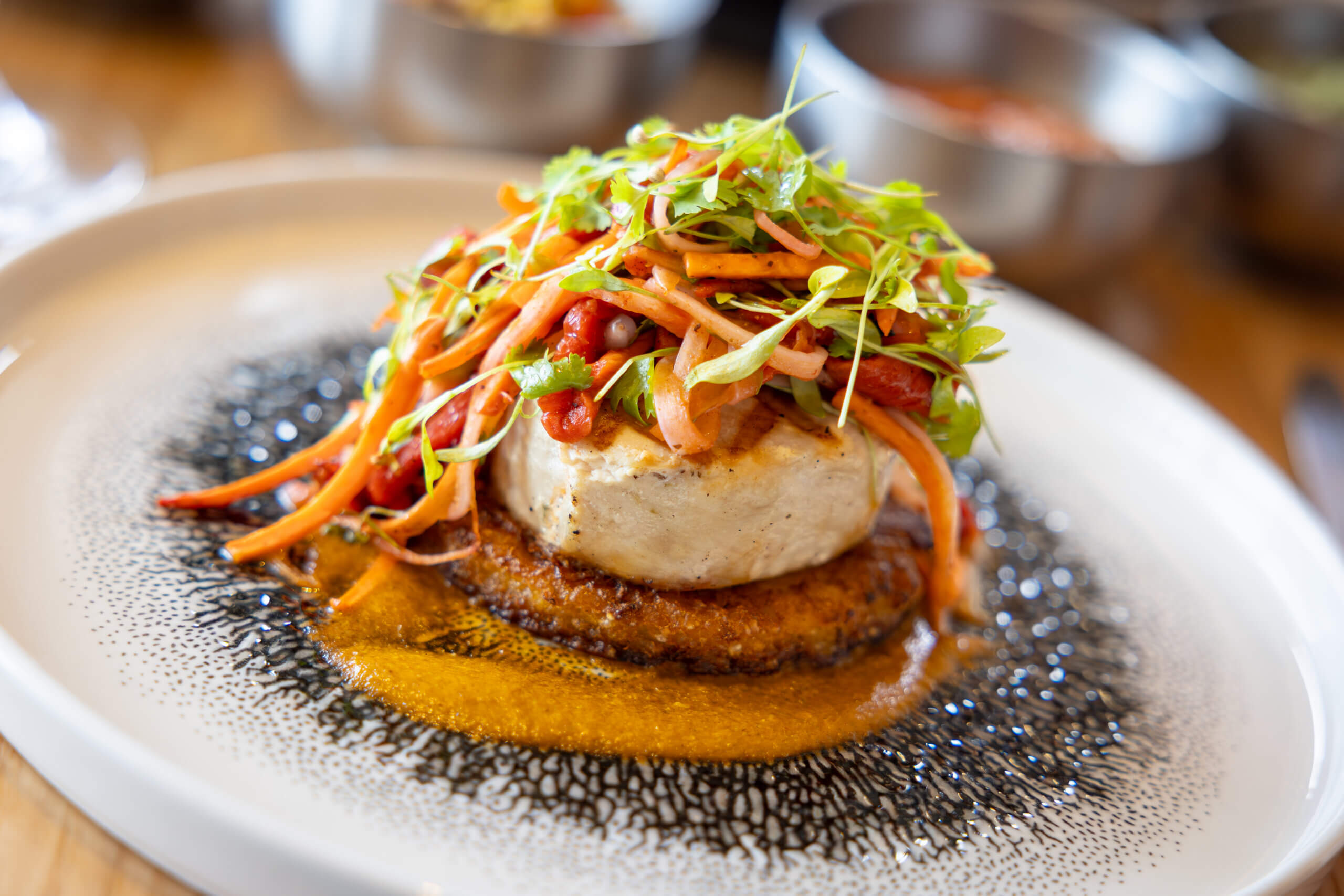 A gourmet dish featuring a seared fish fillet on a crispy base, topped with a colorful julienned vegetable salad and microgreens, served with a rich orange sauce on a modern white plate.