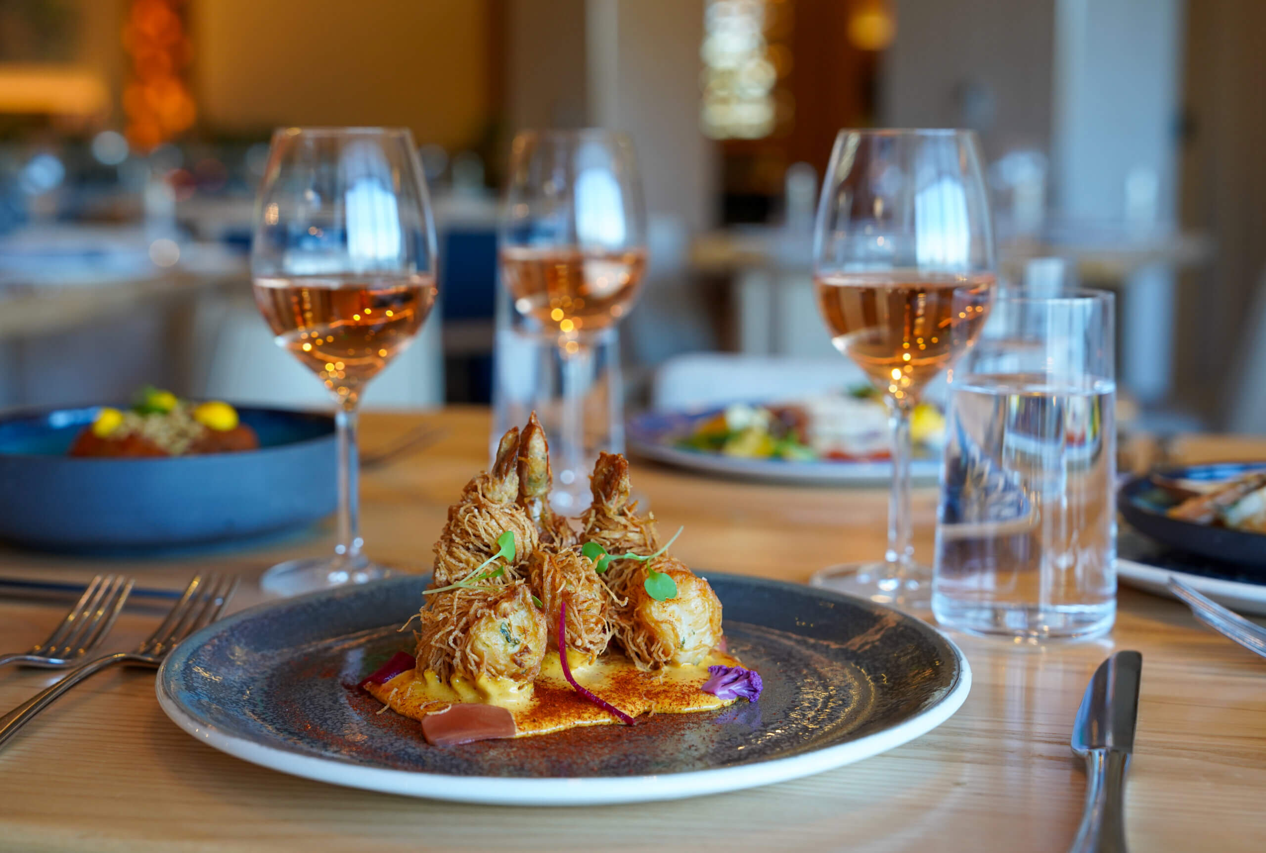 A close-up of a gourmet dish with fried shrimp, garnished with herbs, served on a ceramic plate. Three glasses of rosé wine and a glass of water are on the table, with a blurred restaurant background.