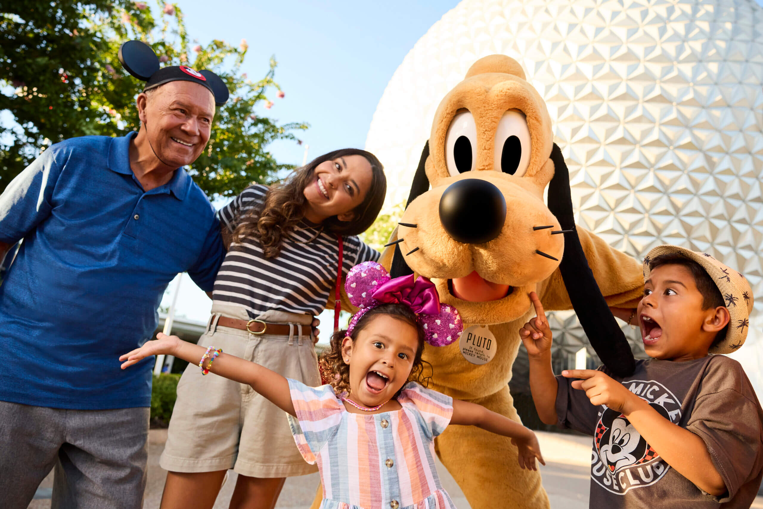 A family smiles and poses with Pluto at a theme park. A large white geodesic dome is in the background. The kids look excited, and everyone is enjoying the moment.