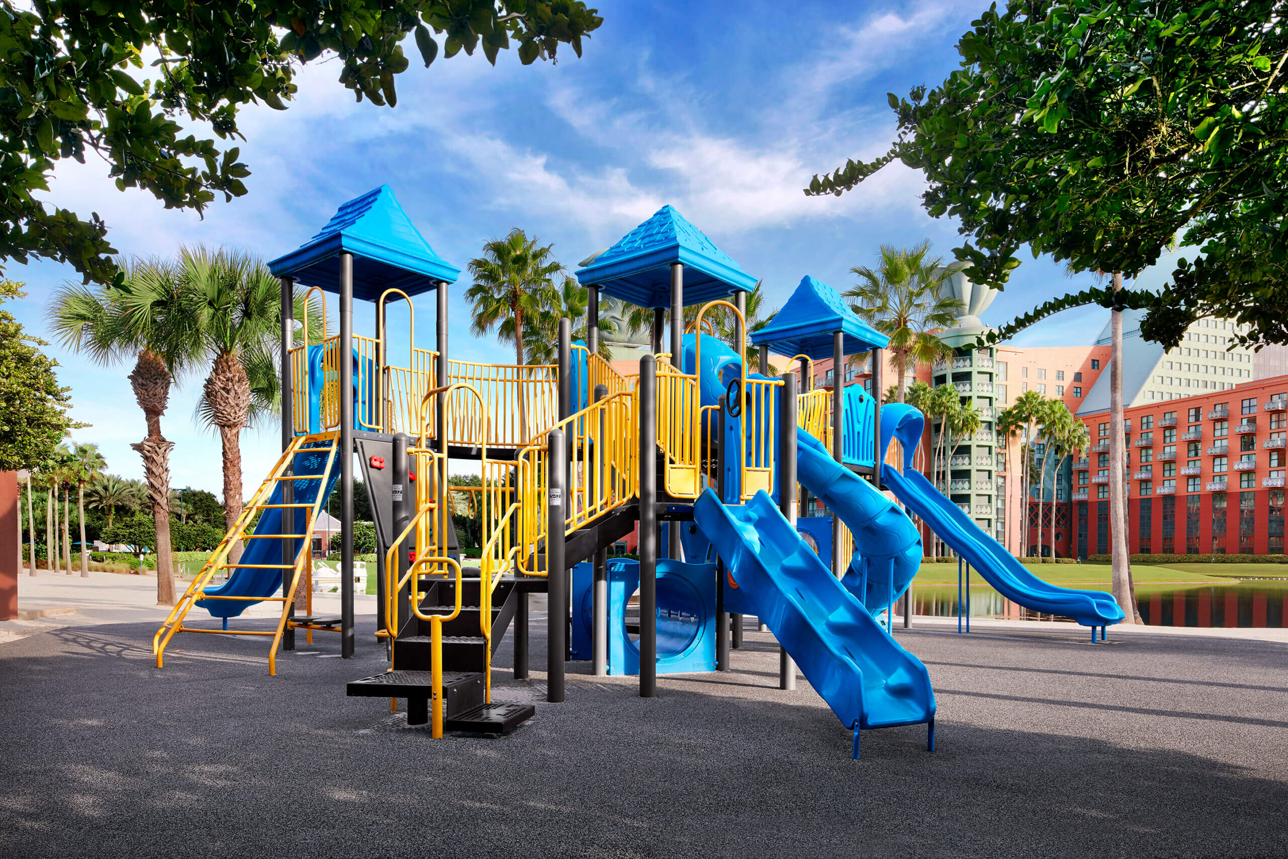 Colorful playground with blue slides, yellow railings, and climbing structures, set outdoors with palm trees and tall buildings in the background under a bright blue sky.