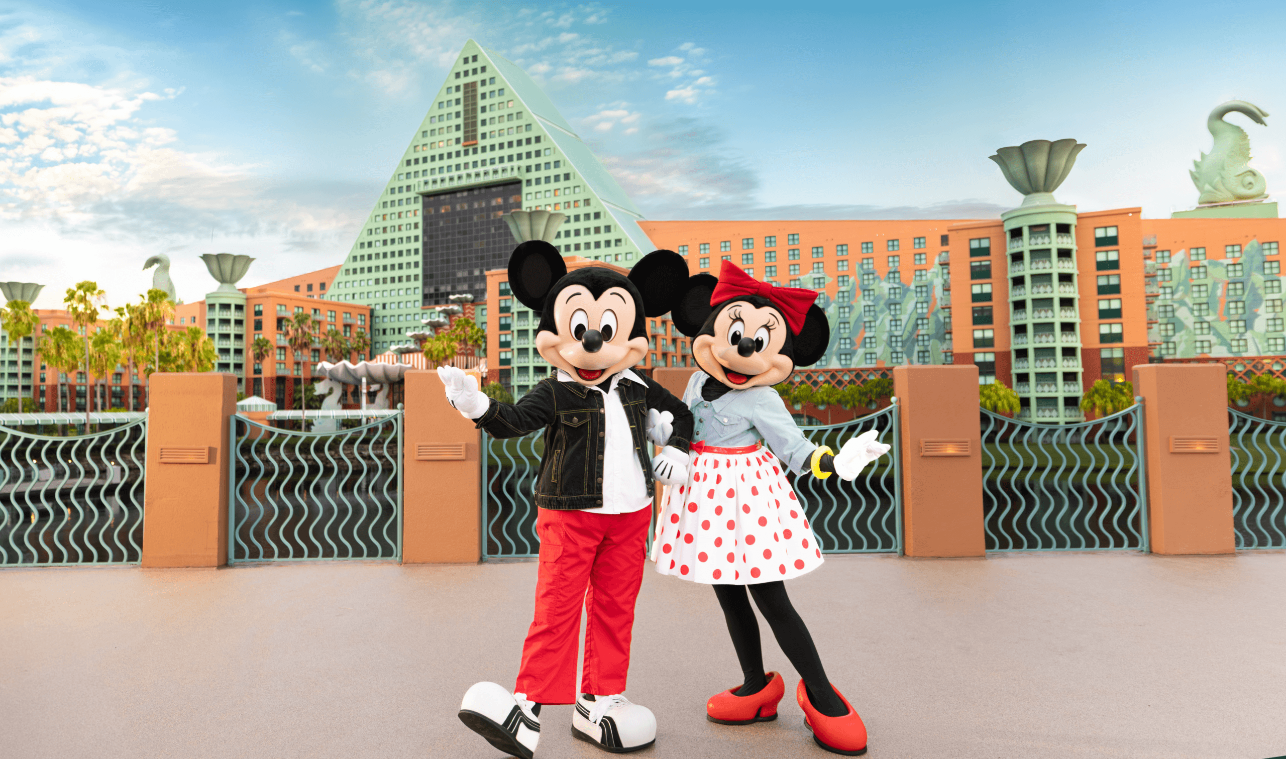 Mickey Mouse and Minnie Mouse pose together in front of the Walt Disney World Dolphin Hotel, with palm trees and blue sky in the background.