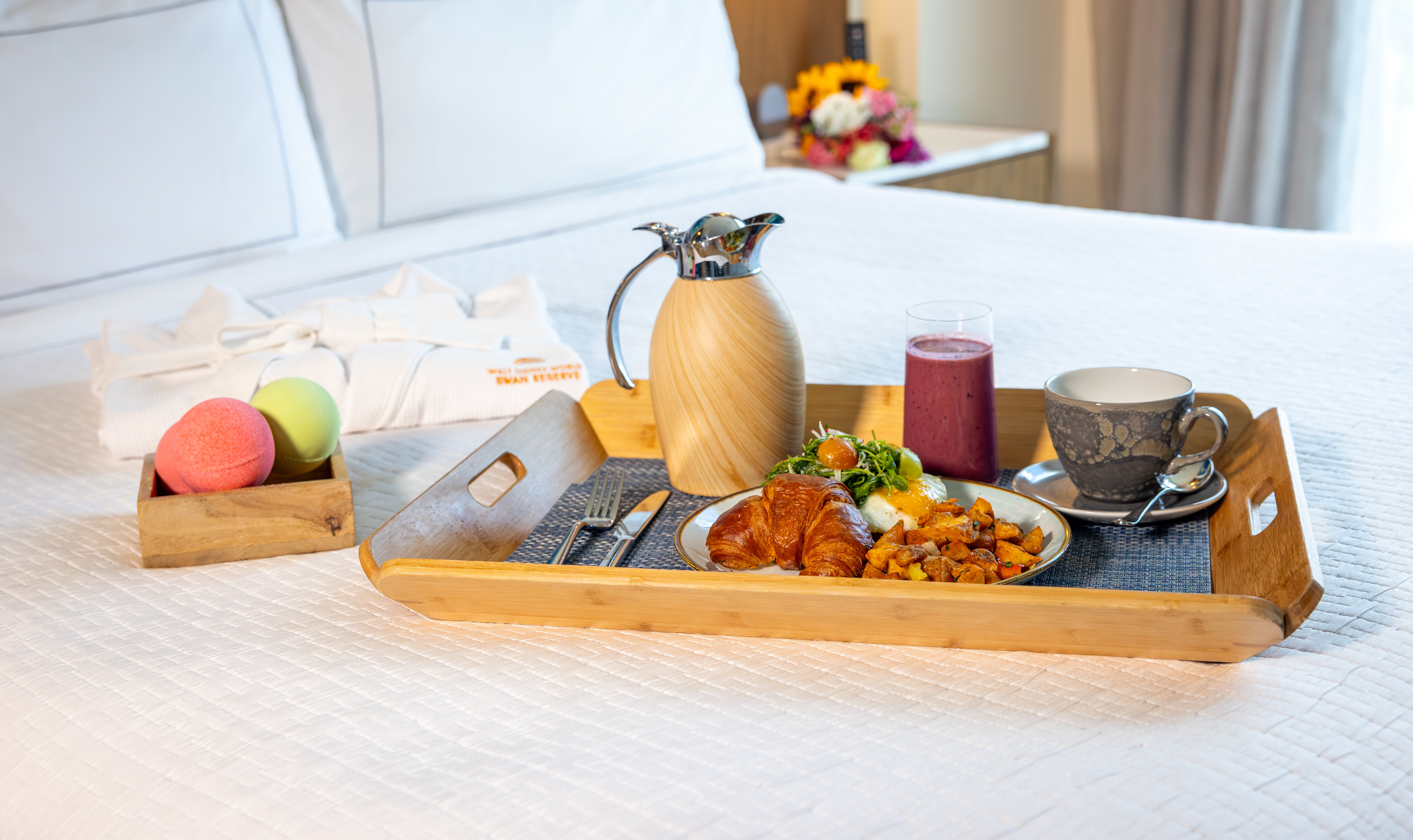 Hotel room service breakfast tray on white bed with croissant, eggs, potatoes, smoothie, coffee, and carafe. Bath bombs and robe nearby; flowers in background.