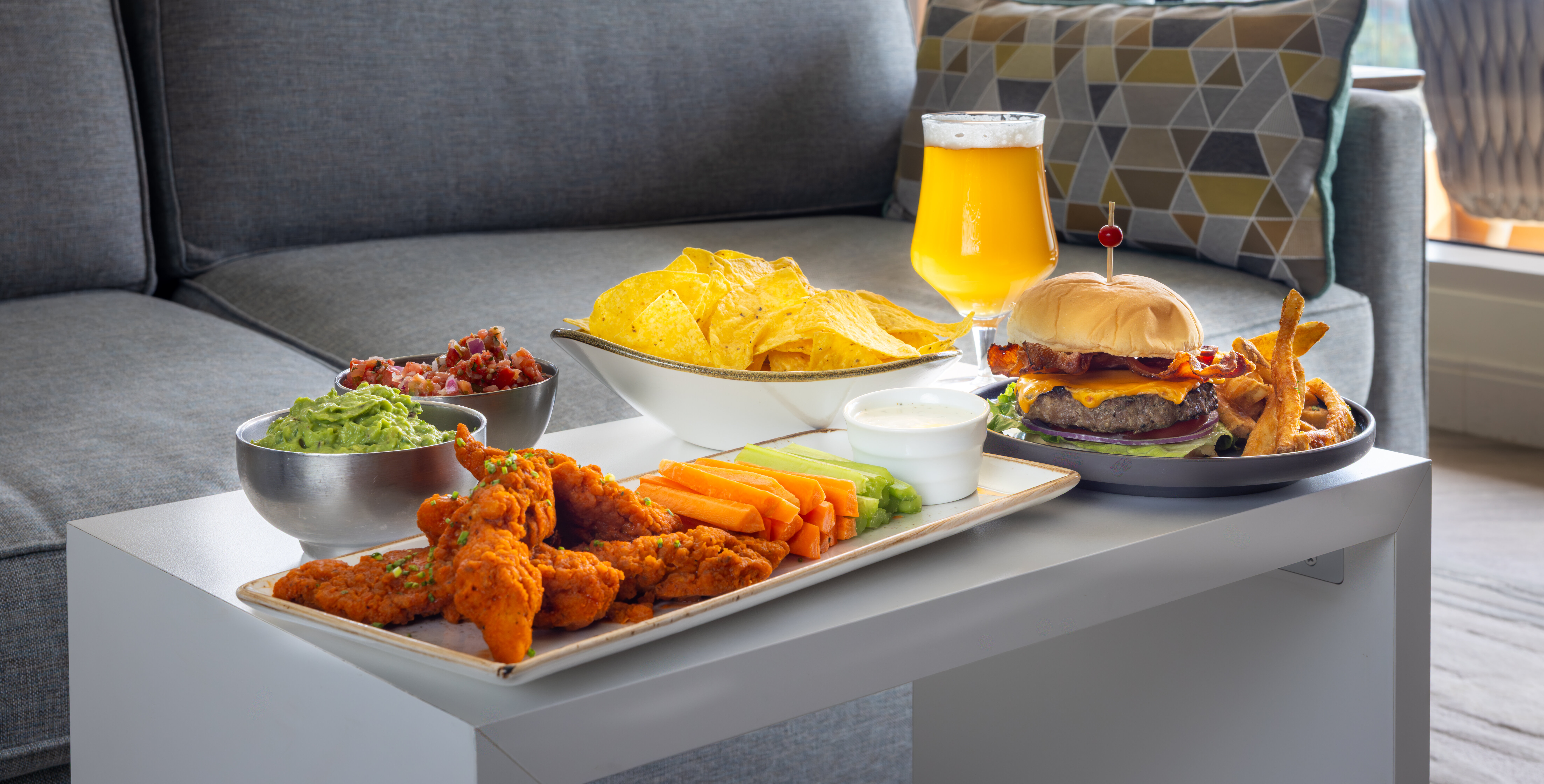A white table holds buffalo wings with veggies and dip, guacamole, salsa, tortilla chips, a burger with fries, and a glass of orange drink in front of a gray sofa in a modern living room.