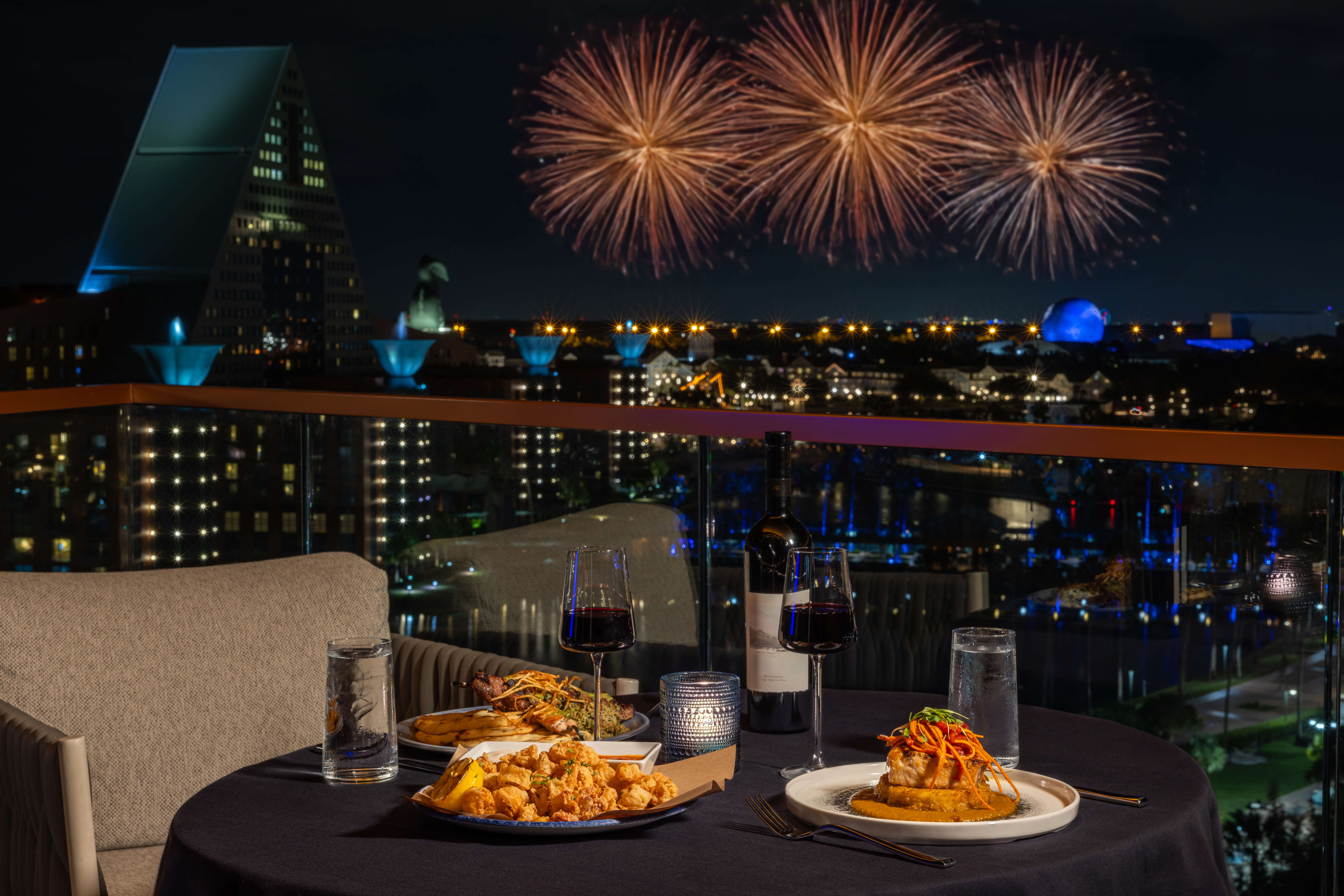 A table on a balcony with food and drinks, overlooking the Dolphin with fireworks in the background.