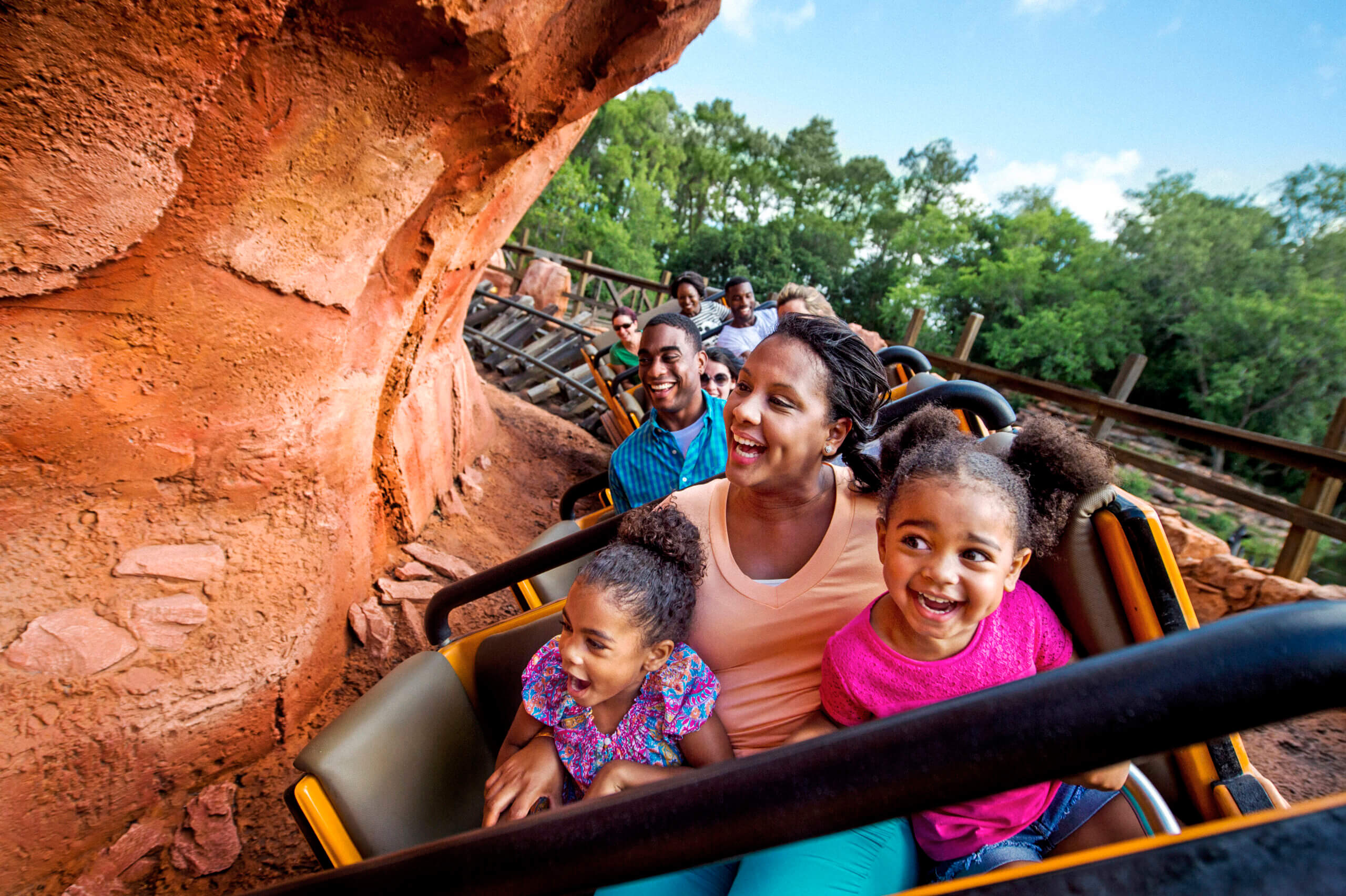 A family with two children smiles and laughs while riding a roller coaster outdoors, surrounded by rocky scenery and trees.
